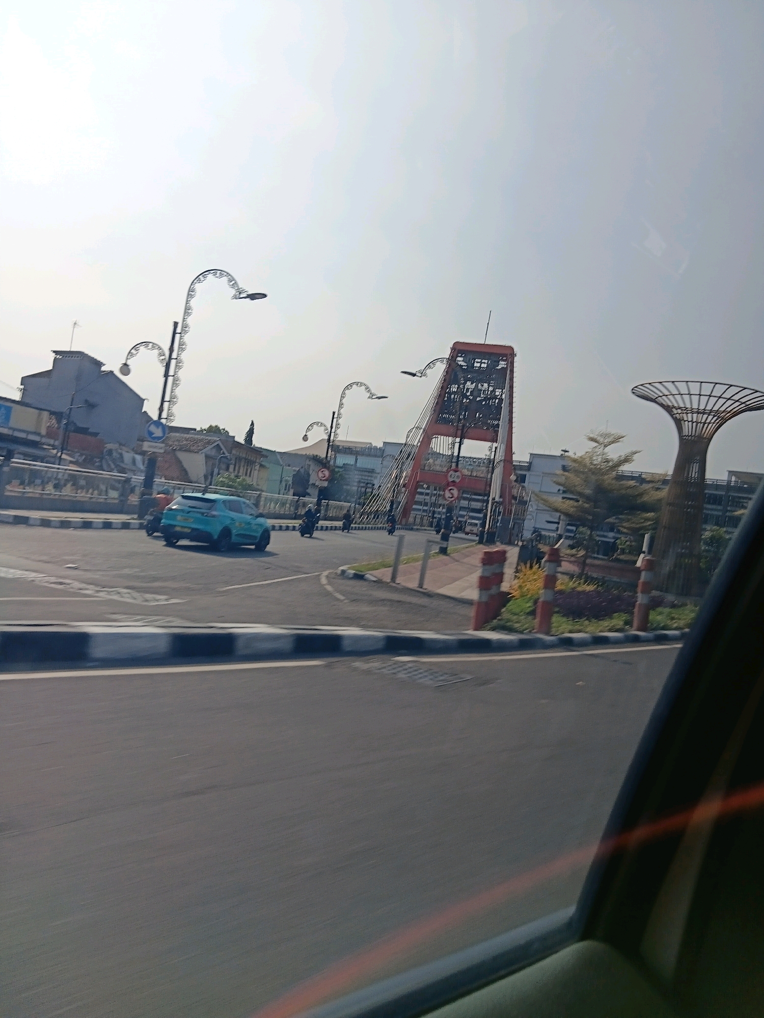A bright, sunny day is captured from the perspective of a vehicle moving through a city intersection in Surabaya, Indonesia. The scene is dominated by the city's infrastructure and a distinctive modern monument.
In the center of the frame, a light blue SUV is turning left at an intersection. It is followed by at least one motorcycle. The road is paved and marked with black and white stripes along the sides, indicating a pedestrian walkway or divider. Traffic lights and street lamps, some with decorative, curved arms, are visible.
Further into the background, a prominent red and white suspension bridge tower rises, its intricate metalwork visible against the pale sky. To the right of the bridge tower, a large, stylized tree-shaped sculpture stands, constructed from metal framing. Buildings of varying architectural styles line the streets in the distance.
The sky is mostly clear with a slight haze. The overall impression is of a busy urban environment on a clear day. The location is identified as Surabaya, Indonesia, likely near a notable landmark.