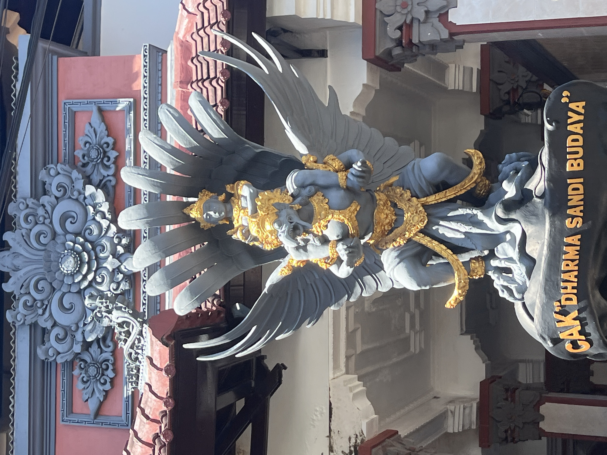 The image shows a large, grey stone sculpture of Garuda, a mythical bird-like creature from Hindu mythology, adorned with gold accents. Garuda is depicted with large wings spread out, carrying a smaller figure on its shoulders. The sculpture is situated in front of a building with ornate grey stonework and a red tiled roof, typical of Balinese architecture. Below the sculpture, a dark stone base bears the inscription "CAK 'DHARMA SANDI BUDAYA'" in gold lettering. The setting appears to be an outdoor cultural site or temple in Kuta Selatan, Indonesia, characterized by bright sunlight and traditional decorative elements.