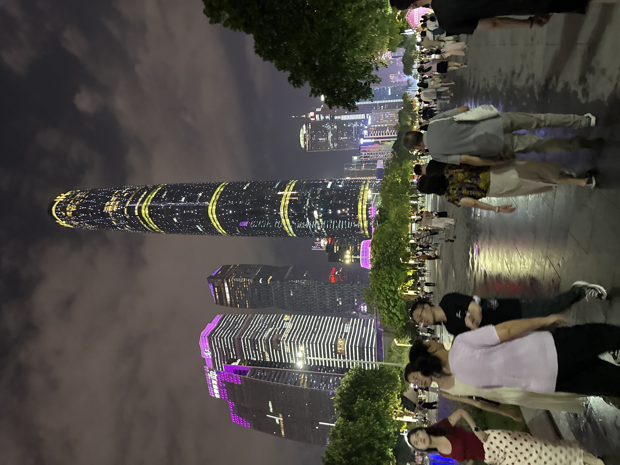 This media file captures a nighttime scene in Tianhe District, China, characterized by illuminated skyscrapers and a gathering of people. The most prominent object is a tall, cylindrical skyscraper with a distinctive design, wrapped in bands of yellow and green lights. Other modern buildings, some adorned with purple and white lighting, punctuate the cityscape.
A group of people are seen walking and standing along a wet pathway, their reflections visible on the slick surface, suggesting recent rain. The overall atmosphere is one of urban nighttime activity, with the bright lights of the city creating a vibrant backdrop. The faces of some individuals are visible, showing a mix of expressions that don't clearly convey a specific emotion, but rather a general engagement with the surroundings.
The time of day is clearly night, due to the darkness and the artificial lighting of the buildings. The wet ground hints at recent weather conditions. The presence of numerous buildings with distinct architectural styles and lighting suggests a modern, developed urban environment. Some text is visible on the buildings, but it is not legible enough to be identified. The activity appears to be casual, with people enjoying the illuminated urban landscape.