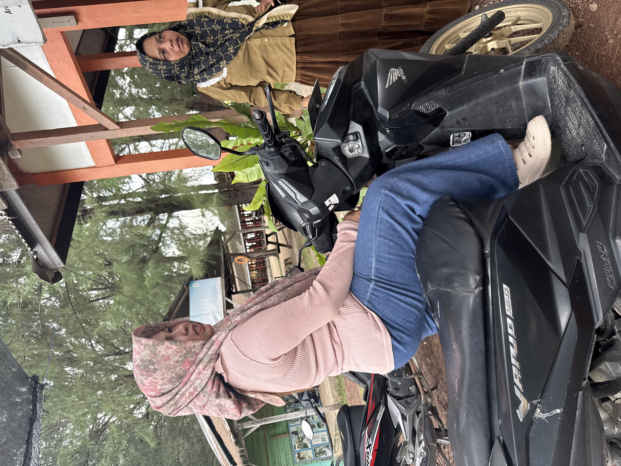 In the city of Meunasahbale, Indonesia, two women are present near a beachside establishment. The woman in the foreground, wearing a pink floral hijab and a pink long-sleeved shirt, is seated on a black scooter. She is also wearing blue jeans and light-colored clogs. The scooter displays the "VARIO 150" logo. In the background, another woman stands, wearing a brown hijab with a black and gold pattern and a brown tiered skirt. She is wearing a brown long-sleeved shirt. The environment suggests a tropical setting with pine trees visible in the background. A sign for "RE CAFE" is partially visible, indicating the presence of a cafe.