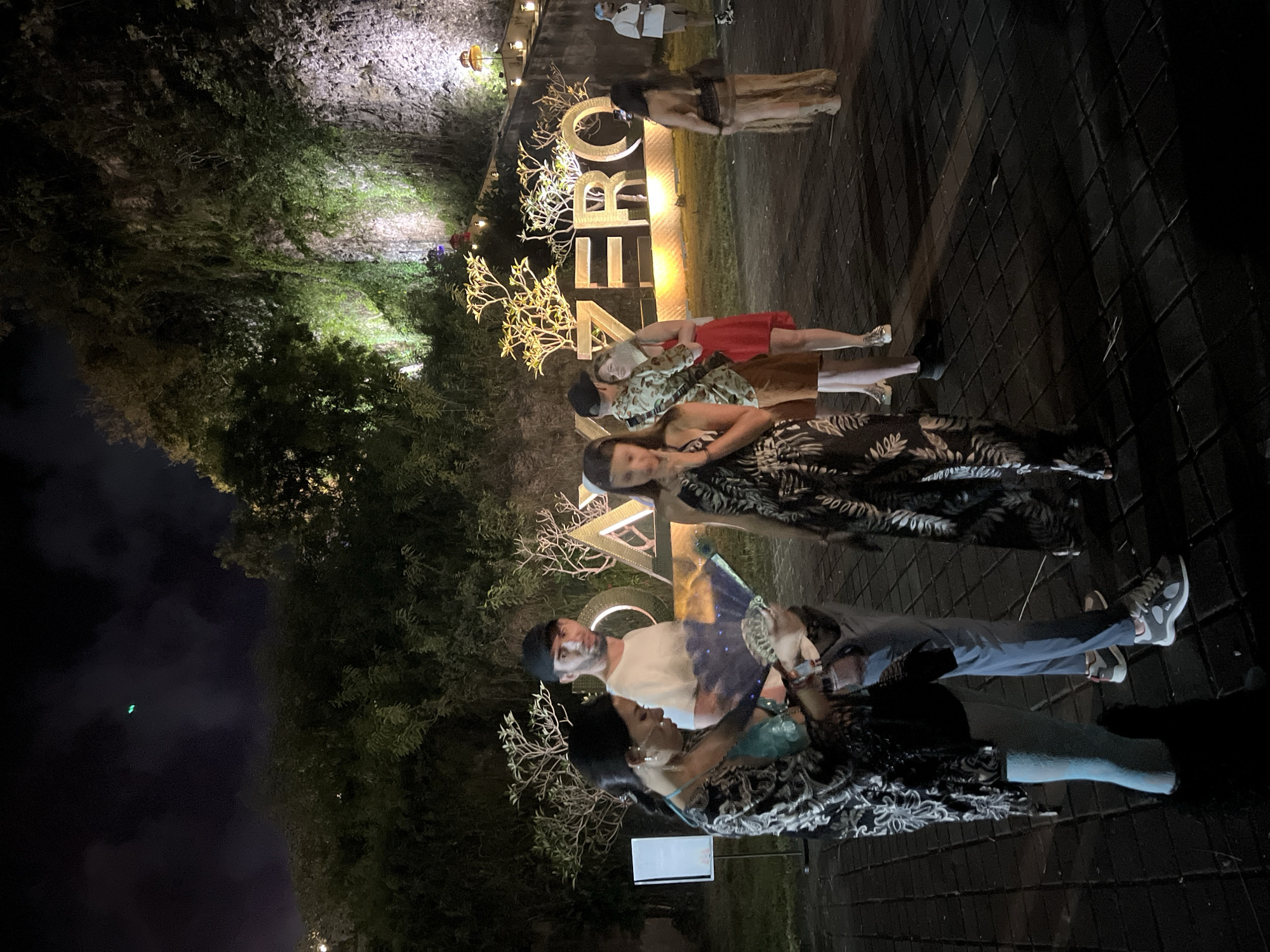 The image captures an outdoor night scene featuring several people gathered around a large, illuminated sign. The setting appears to be a public or event space, with lush, dark green foliage and trees on the left and a textured stone wall on the right. The ground is paved with dark tiles arranged in a diamond pattern.
In the mid-ground, a man with a beard, wearing a white t-shirt, dark pants, and a dark cap, stands next to a woman in a dark, patterned dress who is holding a decorative fan. Behind them, another woman in a similar patterned dress is visible. Further back, a woman in a red dress stands near the central feature of the scene: a large, brightly lit sign spelling out "ZENROB..." in stylized capital letters. The letters are light-colored and have intricate, tree-like decorative elements branching from them, especially around the 'O'. Other individuals are partially visible further in the background, including one person in a white top near the stone wall.
The activity appears to be a casual gathering or photo opportunity, as people are standing and posing near the prominent sign. The time of day is clearly nighttime, as indicated by the darkness and the reliance on artificial lighting from the sign, spotlights on the foliage, and ambient light sources. The expressions on the faces of the visible individuals are mostly neutral or pleasantly calm. The location cues, including the specific sign and the tropical-like foliage, suggest a resort, restaurant, or themed outdoor venue.