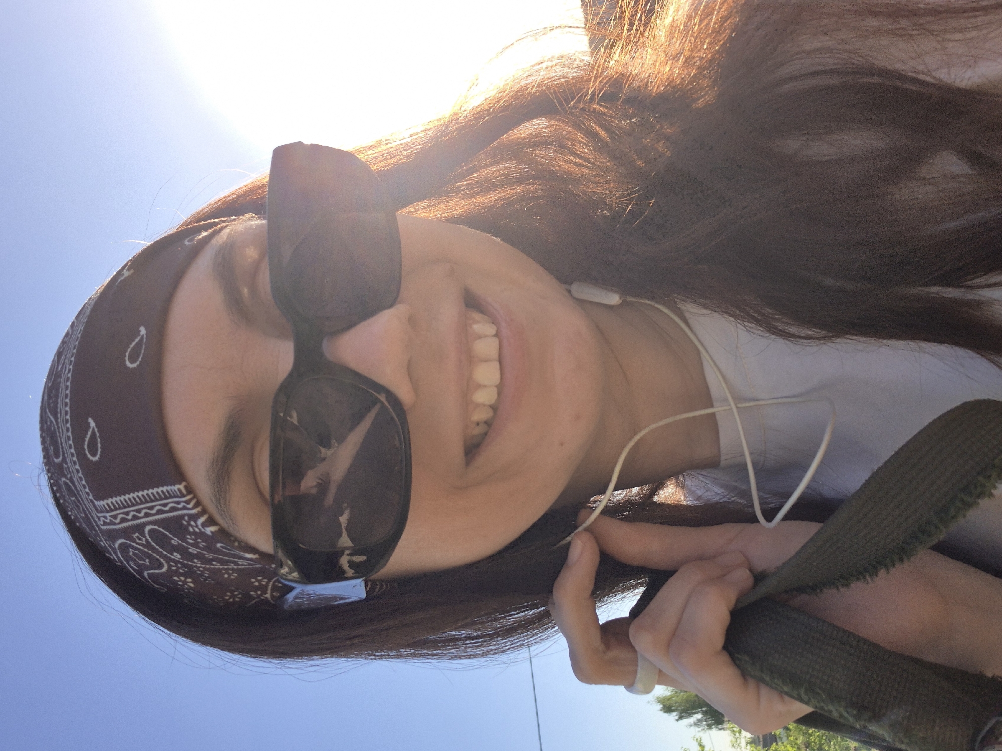 A person is smiling and wearing sunglasses, a bandana, and earphones. They are holding a dark green fabric strap in their hand, and the background shows a bright sky and some greenery. The exact location is unknown.