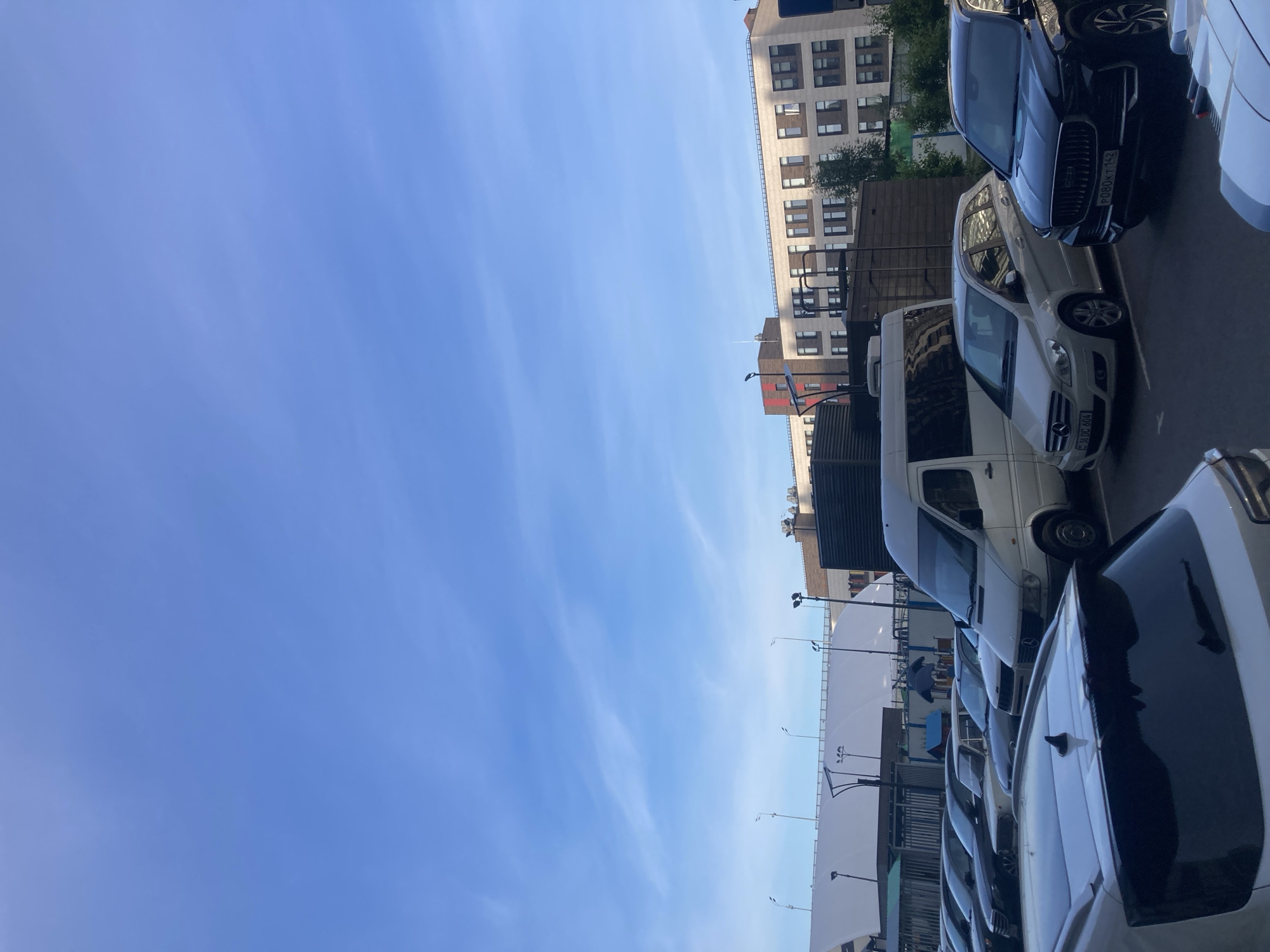 The image, presented in a vertical orientation, captures an urban outdoor scene primarily divided between a clear blue sky on the left and a street or parking area with buildings and vehicles on the right. The sky is largely cloudless, save for a few faint, wispy cirrus clouds.
On the right side of the frame, several vehicles are parked in a line. In the closest foreground, the rear section of a white car with a dark, possibly black, roof or trunk is visible. Behind it, a white Mercedes-Benz commercial van, likely a Sprinter, is clearly seen. Following the van is a light-colored, possibly beige, Mercedes-Benz sedan, which displays a Russian license plate with the characters "F 380 DC 404." Further down the line, a dark-colored SUV, identifiable by its distinctive front grille and headlights, is parked. This SUV also has a Russian license plate, "O80AK 162." Additional cars are partially visible further along the row.
In the mid-ground and background, an urban environment unfolds. A large, multi-story building with a light-colored facade and numerous windows stretches across the background. Another, smaller building with reddish-brown elements is located closer to the street level. Green foliage from trees or shrubs is visible between the buildings and the parked vehicles. Several streetlights or outdoor lamps on poles are present, along with large white tent-like structures or awnings, possibly covering parts of the parking area or an outdoor space.
The scene is set during the daytime, indicated by the bright, overhead lighting and clear sky. The Russian license plates, with region codes "162" and "404" (commonly associated with Russia, with 404 specifically indicating Moscow), affirm the location context of Moscow, Russia. The image is a static snapshot, showing no active movement or people, and conveys a neutral atmosphere typical of an urban street.