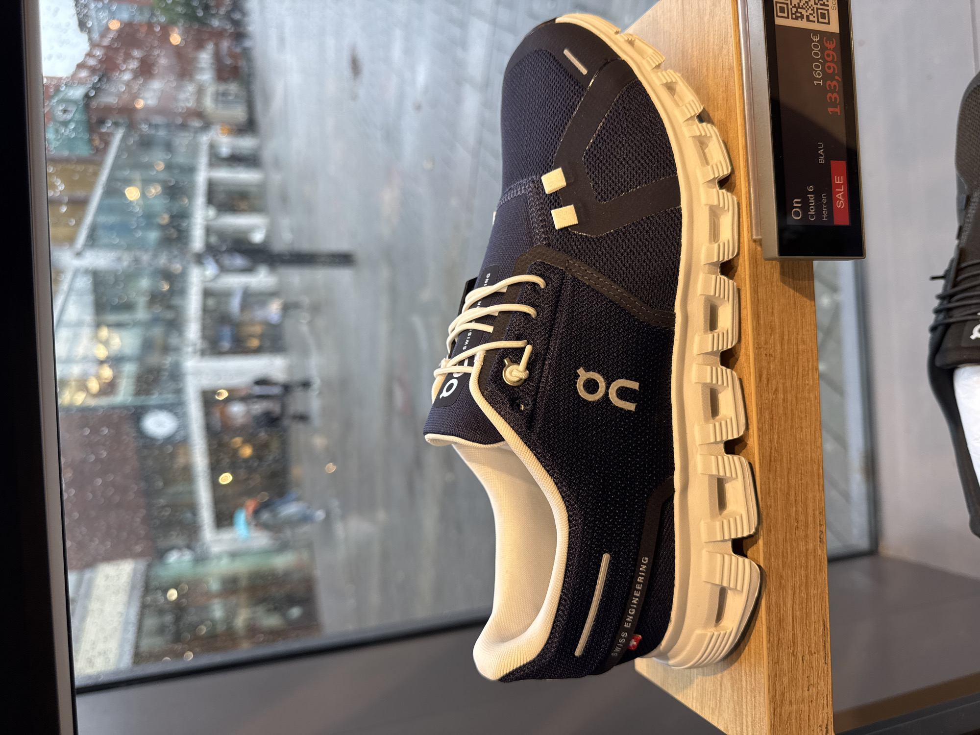 This is a close-up, eye-level shot of a navy blue and white "On Cloud 6" running shoe displayed on a wooden shelf in a retail store. The shoe is positioned with its sole facing the viewer and is angled slightly to the right. The "On" logo is visible on the side of the shoe in white. The shoe has white laces and a white sole with distinctive "cloud" technology. A price tag in the lower right corner displays "On Cloud 6 Herren Blau" with a sale price of "133,99€" and a regular price of "160,00€". In the background, visible through a rain-streaked shop window, is an urban street scene in Münster, Germany. People are seen walking with umbrellas, and buildings with illuminated windows suggest a commercial area. The overall lighting is dim, consistent with a rainy day.