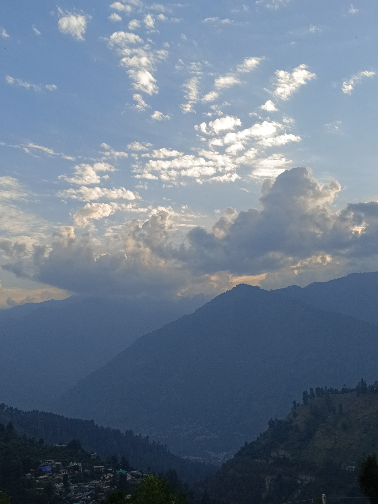 A panoramic view of the mountains in Damadi, India, is captured in this photograph. The sky is a clear blue, dotted with fluffy white clouds that catch the sunlight, creating a dramatic effect. The sun is partially obscured by the clouds, casting a soft glow on the scene. In the foreground, a small village is nestled amidst the rolling hills, with houses of various colors and sizes. Trees line the slopes, adding to the lush green landscape. The mountains in the background are covered in a hazy blue, indicating their distance and the vastness of the landscape. The overall scene evokes a sense of peace and tranquility, with the natural beauty of the mountains and the simplicity of village life.