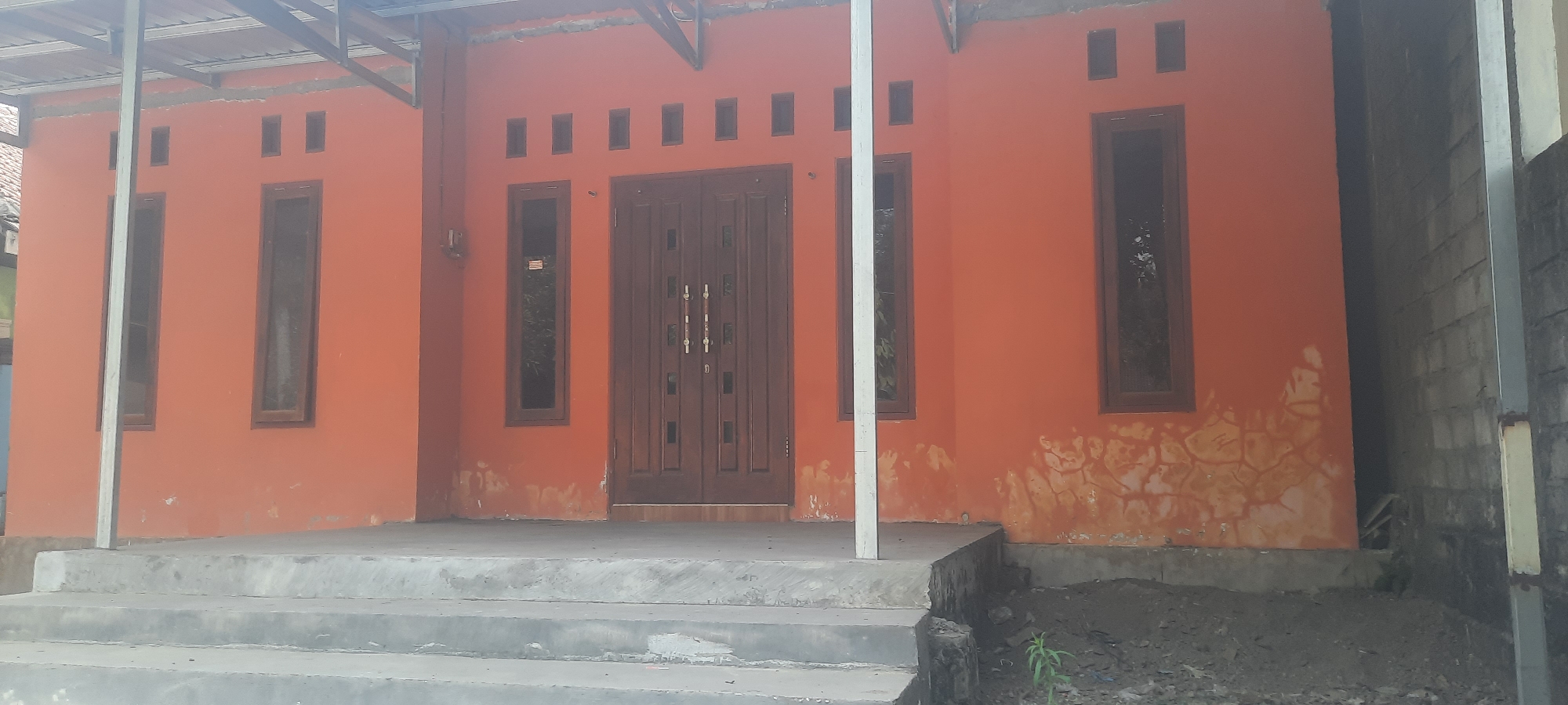 This is a frontal, medium-shot photograph of the exterior of a house. The house is painted orange and features a double door in the center. To the left and right of the door are tall, narrow windows with dark frames. Above the windows and door, there are several small rectangular openings, possibly vents. The house is set back from the street and accessed by a concrete staircase. To the right of the house, there is a gray cinder block wall. The area in front of the house appears to be under construction or landscaping, with piles of dirt. The photograph was taken in Ploso, Indonesia.
