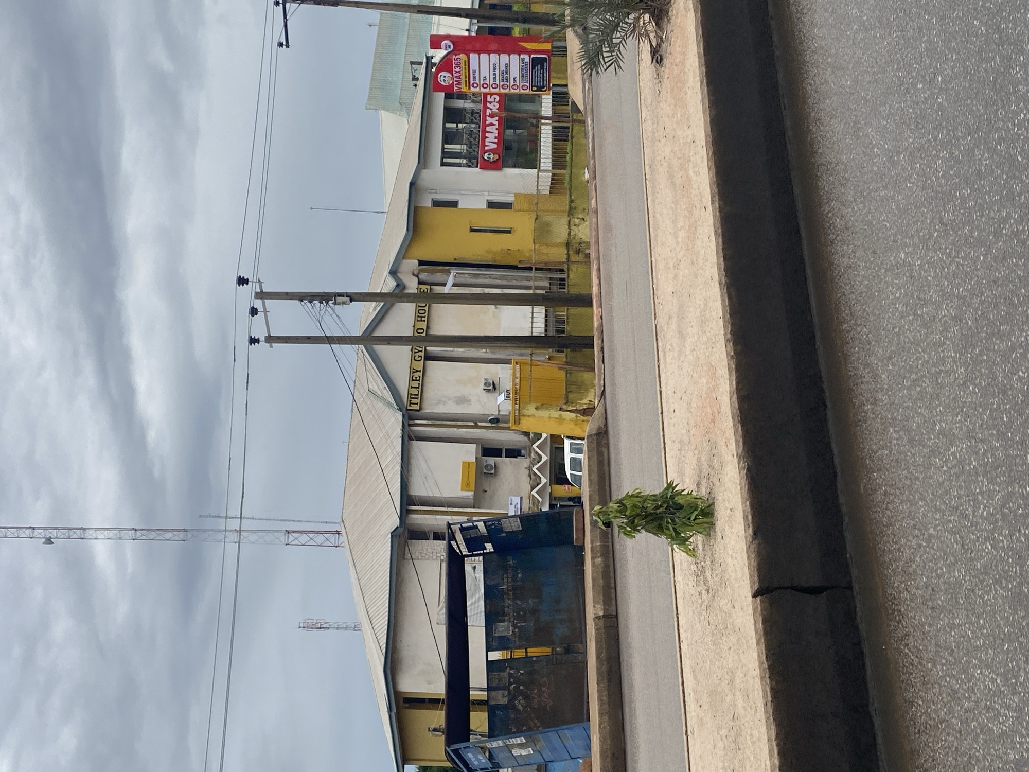 The image depicts a street scene in Jos, Nigeria, under a cloudy sky. In the background, a two-story building with white and yellow sections has the text "TILLEY GYM HO" visible on its facade. A red and white sign for "VMAX365" is prominently displayed on the right side of the building, listing services such as coffee, tea, solid food, snacks, drinks, spa, and aesthetics and accessories. In front of the building, there is a large, blue, dilapidated refuse container. A small, leafy tree is planted on a raised concrete median separating the road from the sidewalk. The road surface shows signs of wear. The environment appears to be urban, with utility poles and wires present.