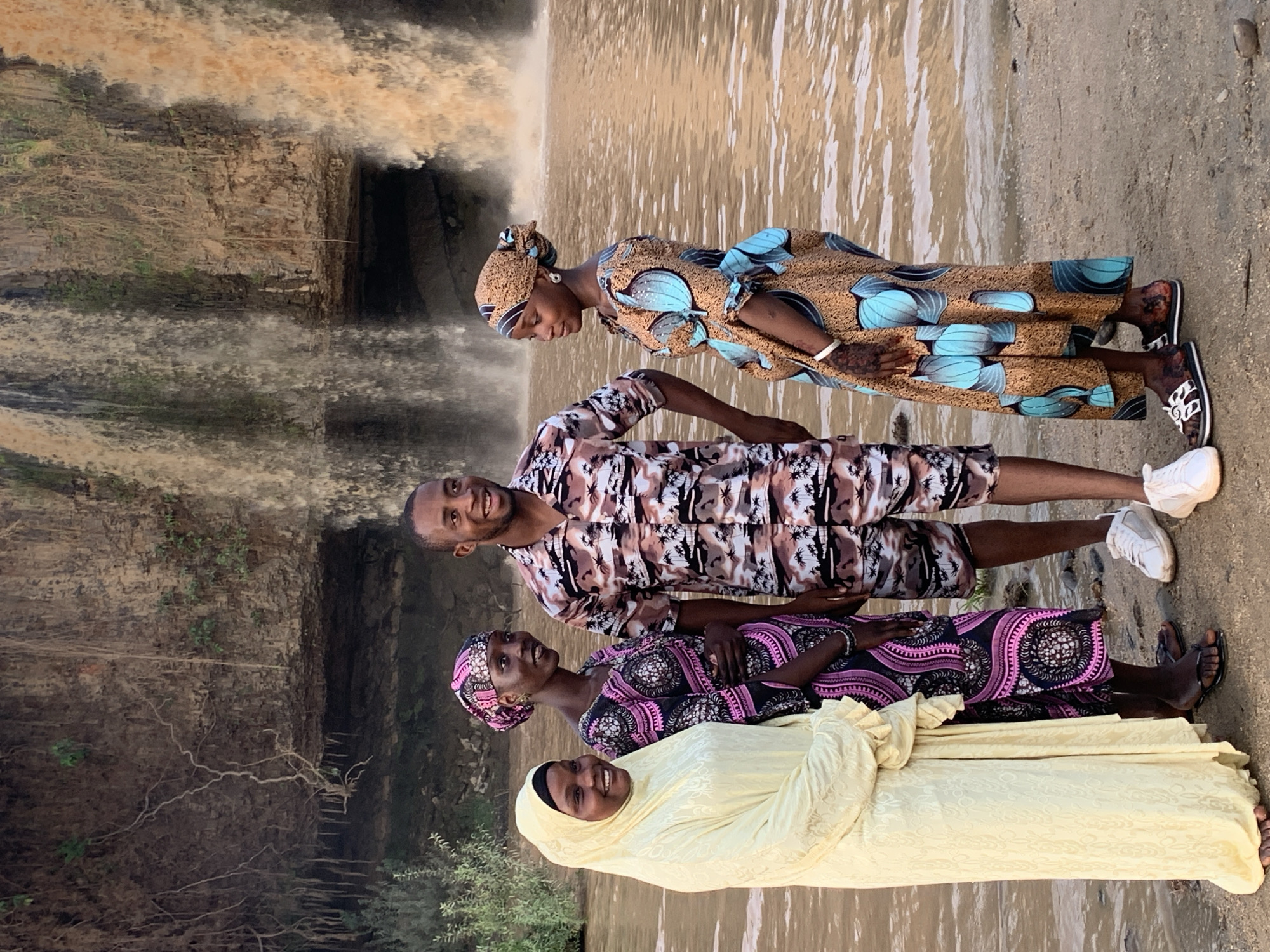 Four people stand on a sandy bank in front of a waterfall. The person closest to the camera wears a light yellow head covering and long dress. A woman in a black and pink patterned dress and head wrap stands next to her. A man stands next to her, wearing a patterned shirt and shorts. Furthest from the camera, a woman in a brown and turquoise patterned dress and head wrap stands. The waterfall is in the background. This scene is in the city of Kukum, Nigeria.