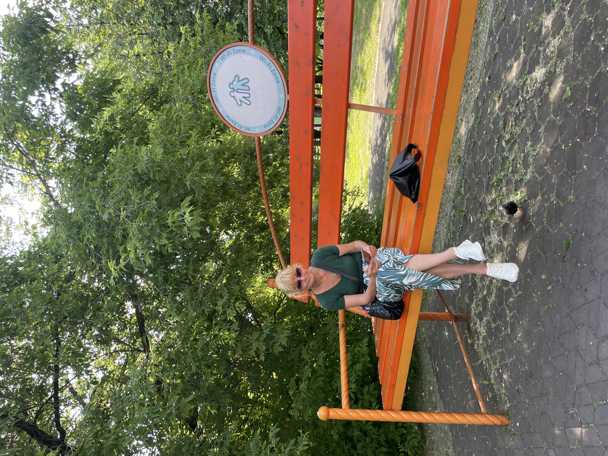 A medium shot captures a blonde woman in sunglasses, a green short-sleeved shirt, and a patterned skirt, sitting on an orange bench in a park. She holds a mobile phone in her hands and has a black purse on the bench beside her. A black pigeon walks on the paved ground in front of the bench. A circular sign with the text "Wi-Fi Zone" and a graphic of two people is visible in the background, attached to a railing above the bench. The scene is set amidst lush green trees and dappled sunlight. The location is the park near the "Zolotaya Riba" (Golden Fish) playground in Novokuznetsk, Russia.