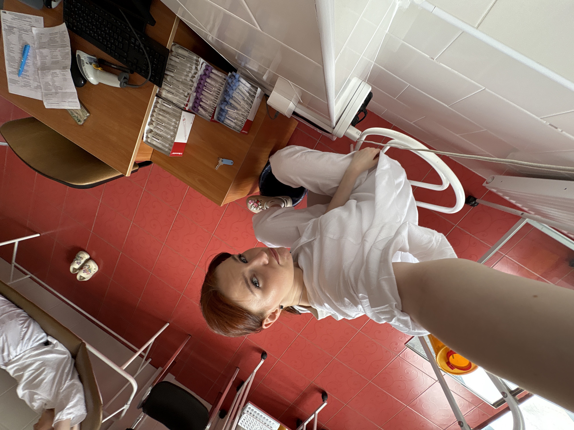 The image shows a person with fair skin and reddish-brown hair, wearing a white shirt and seated on a chair. The location appears to be a medical office, judging from the tiled walls and floor, medical supplies, examination table, and computer equipment visible in the background. The person seems to be taking a selfie. The exact location within Saint Petersburg, Russia, cannot be determined from the visual data.