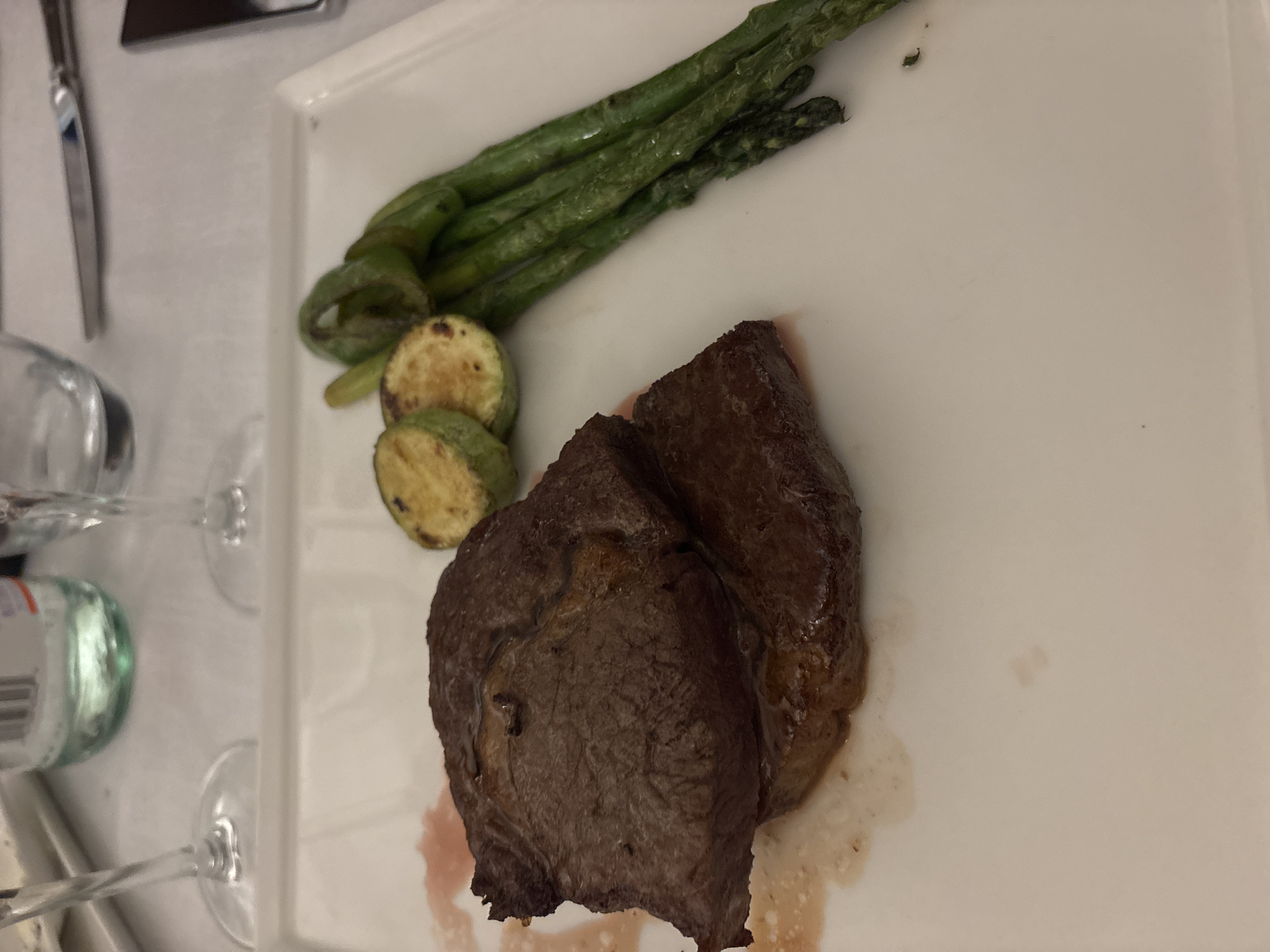 A plated meal of two steak medallions, sliced zucchini, and asparagus is presented on a white rectangular plate. The steaks are dark brown and appear to be medium-well to well-done, with some juices on the plate. The zucchini slices are round and have grill marks. The asparagus spears are green and appear cooked. The setting is indoors, likely a restaurant, with a white tablecloth visible. The image does not provide information about the specific location in Varna, Bulgaria.