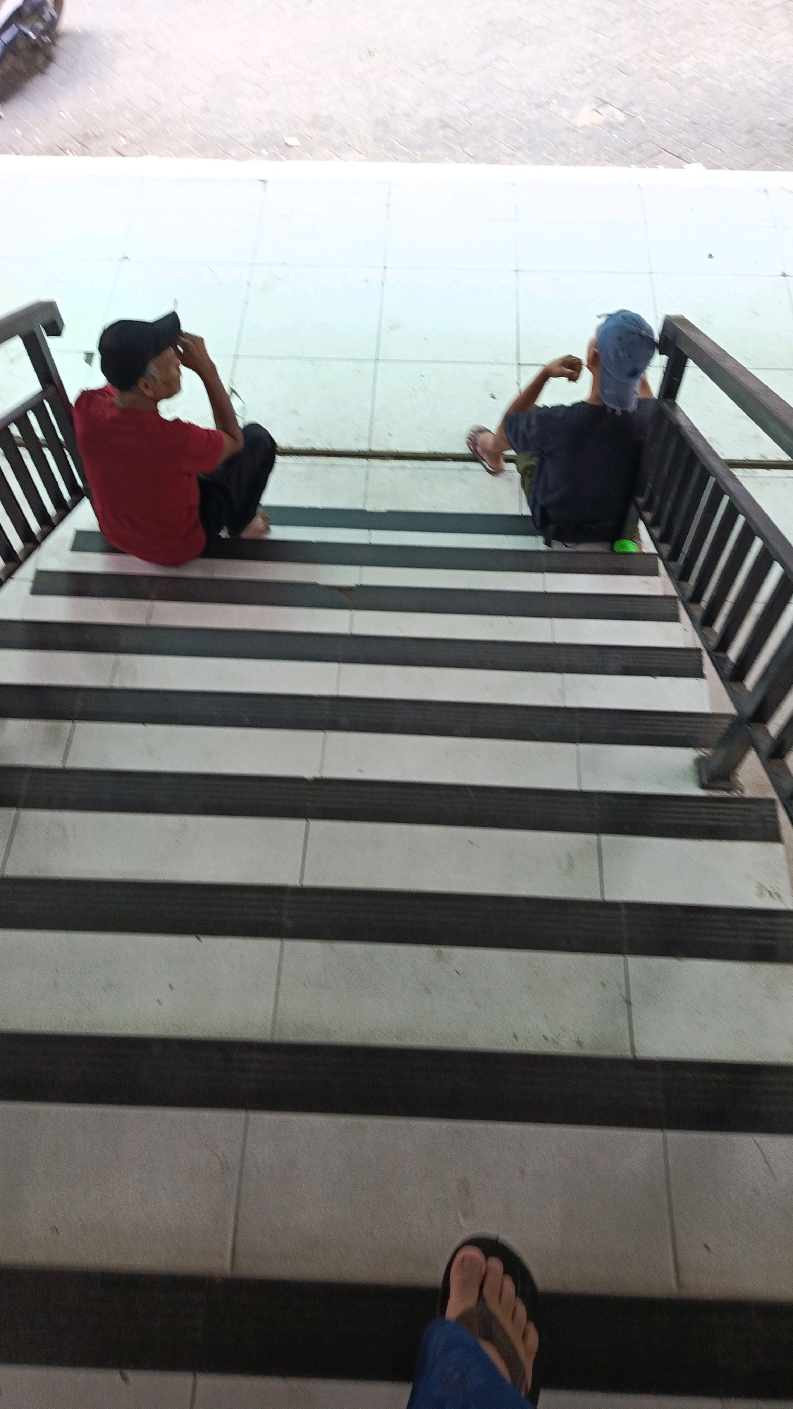 In Ponorogo, Indonesia, two individuals are seated on a set of outdoor stairs. The person on the left, wearing a red t-shirt and a black cap, is looking to the side. To their right, another individual, dressed in a dark t-shirt and a blue cap, is also seated, facing away from the camera. The stairs are characterized by alternating dark and light-colored treads, with a dark railing on the right side of the frame. The ground level above the stairs appears to be paved with white tiles. The perspective is from a higher vantage point looking down the stairs.