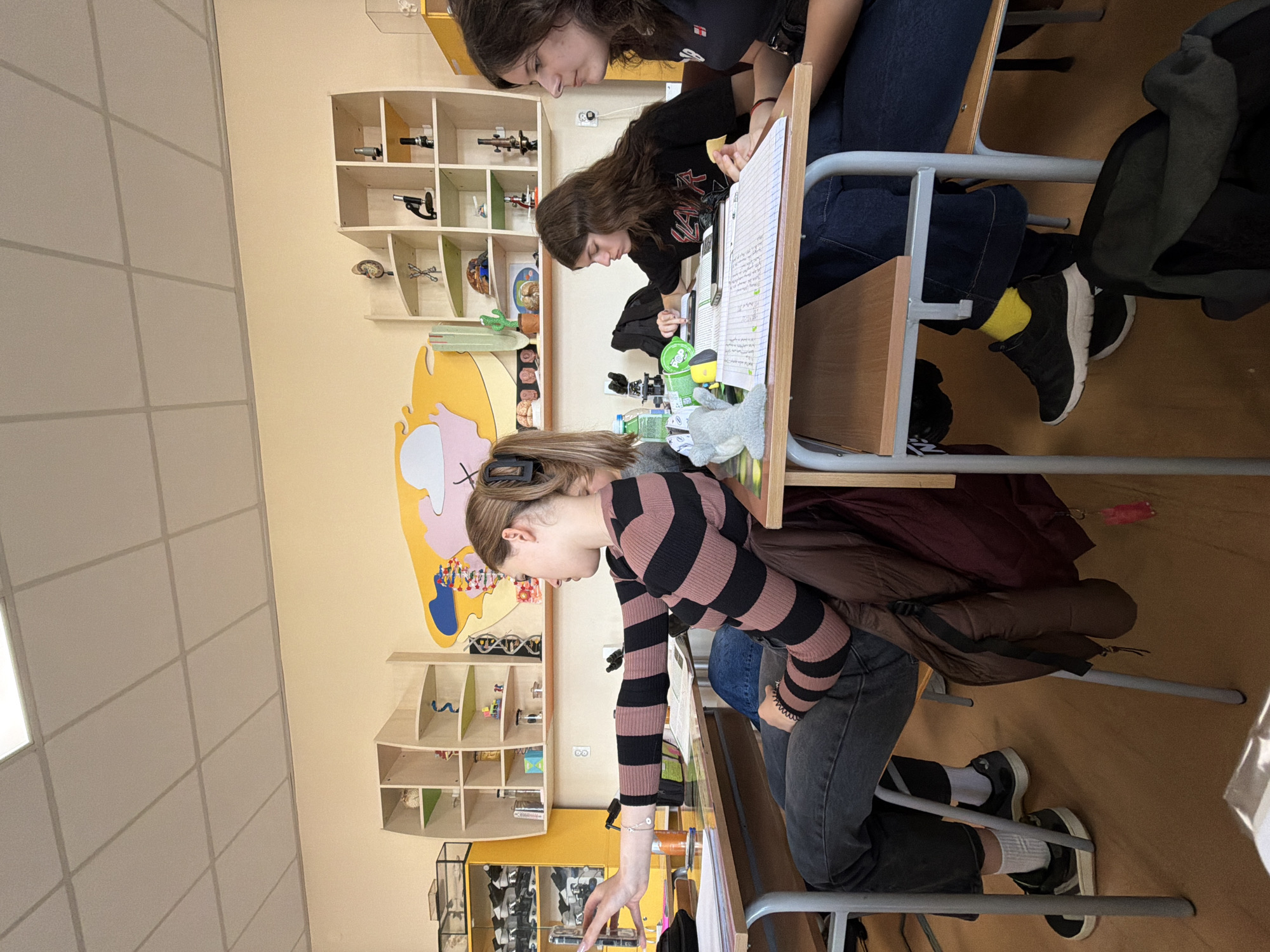 Three individuals are seated at desks in a classroom in Panagyurishte, Bulgaria. They are seemingly engaged in academic work, with one individual focusing intensely on a phone. The classroom contains shelving units displaying models and other objects.