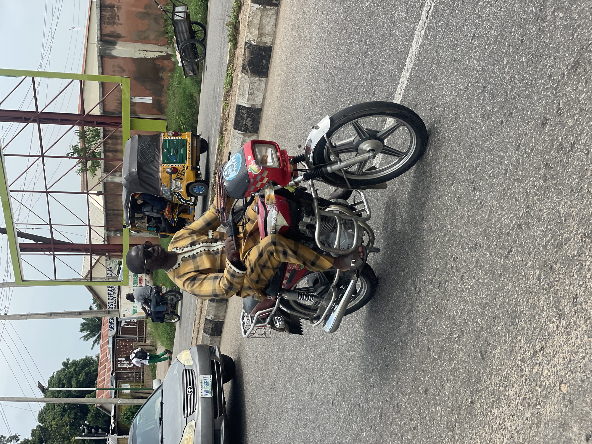 A man wearing a yellow and brown patterned outfit and sunglasses rides a red and silver motorcycle on a paved road in Lokoja, Nigeria. The man is looking forward as he steers the motorcycle, which has a red headlight casing decorated with stickers. The road is lined with a curb that has alternating black and white painted sections. In the background, a grey sedan is visible on the left, and behind it, a building with "GENERAL POST OFFICE LOKOJA" is partially visible. To the right of the motorcycle, a yellow and black auto-rickshaw is moving in the same direction. Further in the distance, a large green billboard structure stands against a cloudy sky. The environment appears to be an urban setting with some greenery and utility poles visible.
