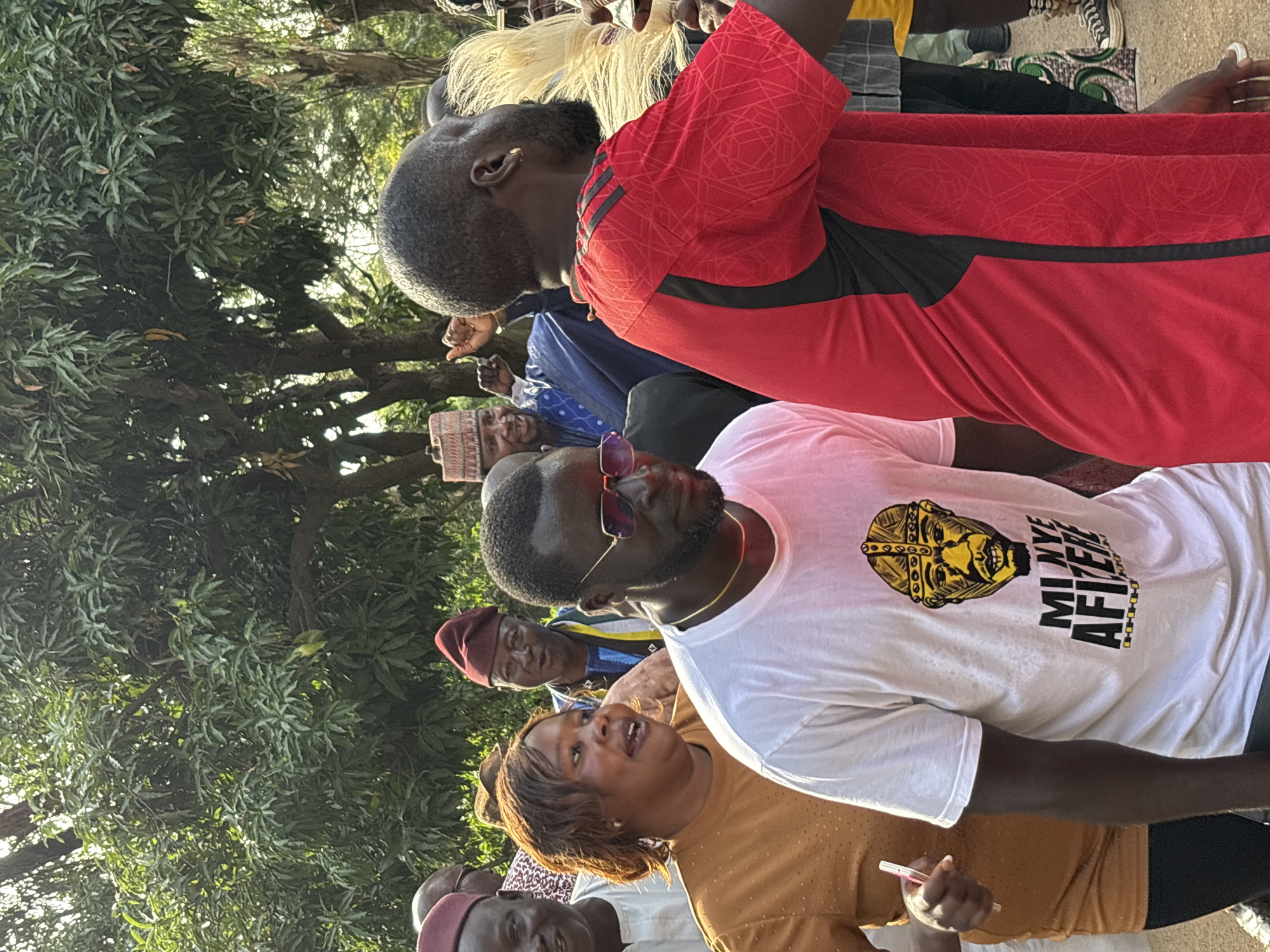 The image shows a group of people outdoors, likely in a public space in Jos, Nigeria, given the context. The lighting suggests it is daytime, possibly late afternoon or early evening due to the warm light. The setting appears to be a street or open area with trees and foliage in the background, indicating an outdoor gathering.
Several individuals are visible, some with their faces turned towards the camera or engaged with others. In the foreground, a woman in a tan top is looking upwards and speaking, her expression lively. To her left, a man in a white t-shirt with a graphic print is looking towards the right. The graphic on his shirt features a portrait of a person with a crown and the text "MI NYE AFTER". To the left of the woman, another person's head wearing a maroon cap is visible. Behind them, a man in a red shirt with black accents is looking down. Further back, a man wearing a blue patterned shirt and a cap is also visible. The people appear to be of African descent, and their clothing suggests a casual or possibly ceremonial gathering.
The overall atmosphere seems to be one of engagement and social interaction. The composition of the photo, with people close together and looking in various directions, suggests a dynamic event or a candid moment captured. The presence of trees and the natural lighting place the scene outdoors.