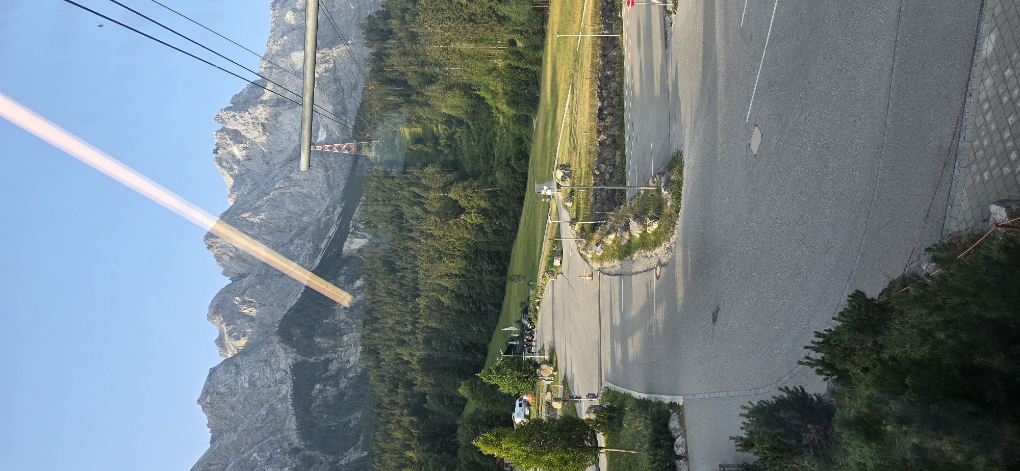 The image captures a mountainous landscape with a road in the foreground, likely near the Zugspitze cable car station in Grainau, Germany. A clear blue sky dominates the upper portion of the image, with a prominent light reflection streaking across it. Rugged, rocky mountains form the backdrop, partially covered with lush green pine forests. A red and white pylon for a cable car system stands out against the mountain slope, with cables extending from it towards the foreground.
In the foreground, a paved road with white lane markings curves through the scene. Several parked vehicles, including a white camper van and a row of motorcycles, are visible near a cluster of trees and buildings on the left side of the road. Rocky outcrops and strategically placed lighting fixtures line the sides of the road and parking areas. The ground is a mix of asphalt and paved stone pathways, with manicured green grass and low-lying bushes interspersed with larger rocks. The overall impression is one of a well-maintained tourist area set against a majestic natural backdrop.