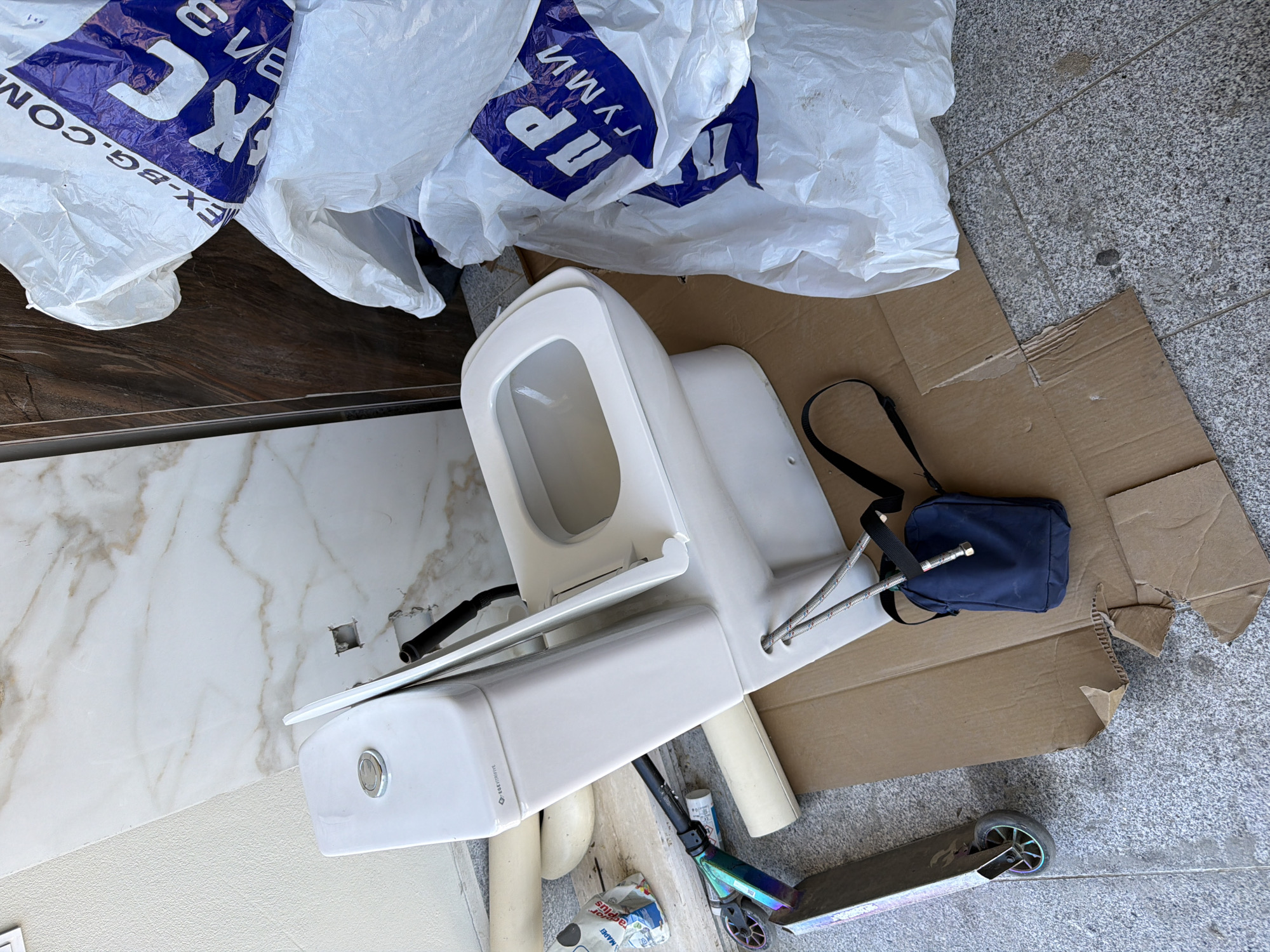 The scene depicts a chaotic assortment of objects on what appears to be an outdoor tiled or concrete surface, likely a sidewalk or patio. Predominantly, a white, disassembled toilet fixture occupies the center of the frame, resting on a piece of brown cardboard. Adjacent to the toilet is a partially visible, multicolored scooter. A navy blue bag with a black strap lies near the toilet, also on the cardboard.
Scattered around the toilet are several plastic bags, some printed with the text "EX-BG.COM" and "ПЪЛНИ" in blue lettering. A bag or container with the word "MAPEI" is also visible.
The background consists of marble-patterned tiles on one side, and gray concrete tiles on the other.
There are no people visible in the image.
The location is not discernible from the image content itself. There is no information included in the image to suggest the city of Asenovgrad, Bulgaria.