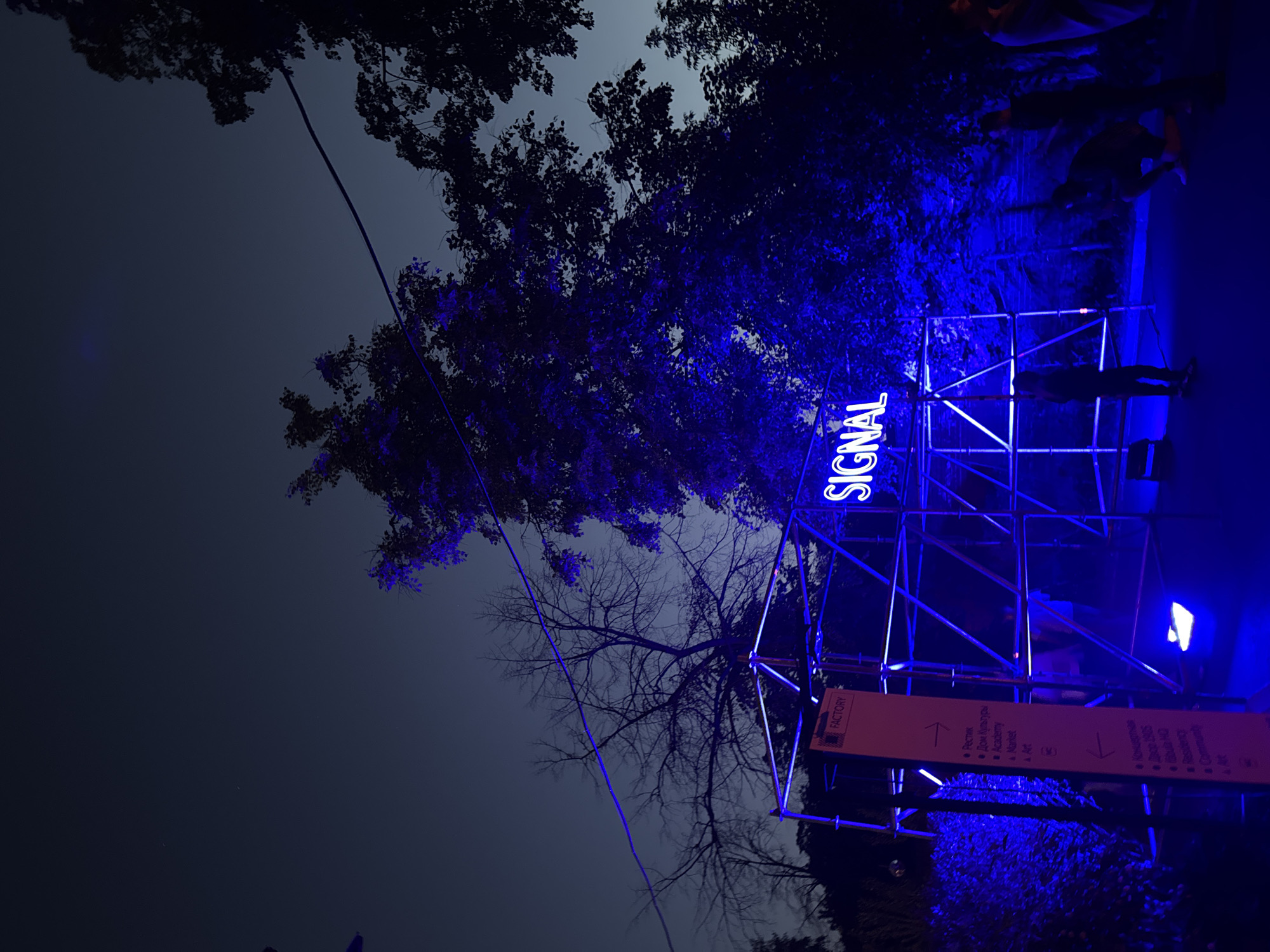 The image depicts a nighttime outdoor scene dominated by blue neon illumination, likely at an arts or cultural event within a creative cluster in the city of Moscow, Russia. A large blue neon sign spells "SIGNAL" and is mounted on a metal scaffolding structure in the center-right foreground. To the left of this structure, an orange directional sign, topped with the word "FACTORY," indicates various venues. A right-pointing arrow directs towards "Restaurant," "House of Culture," "Academy," "Market," and "Art." A left-pointing arrow indicates "Complex," "Yard," "School," "Residency," "Community," and "Art."
The scaffolding itself is also outlined with blue neon lighting, casting a strong blue glow onto the immediate surroundings, including the ground, nearby trees, and people. Several individuals are present: one person stands facing away near the base of the scaffolding to the left, another crouches or sits on the ground to the right of the scaffolding, and additional indistinct figures are visible further right in the background. The people are largely silhouetted by the intense blue light. Trees with dark foliage, some appearing bare, frame the upper and left portions of the image, also touched by the blue glow. An overhead wire crosses the sky from left to right. The sky above is dark blue, characteristic of dusk or night. The ground appears to be a paved pathway. The overall environment suggests an urban or semi-urban artistic venue.