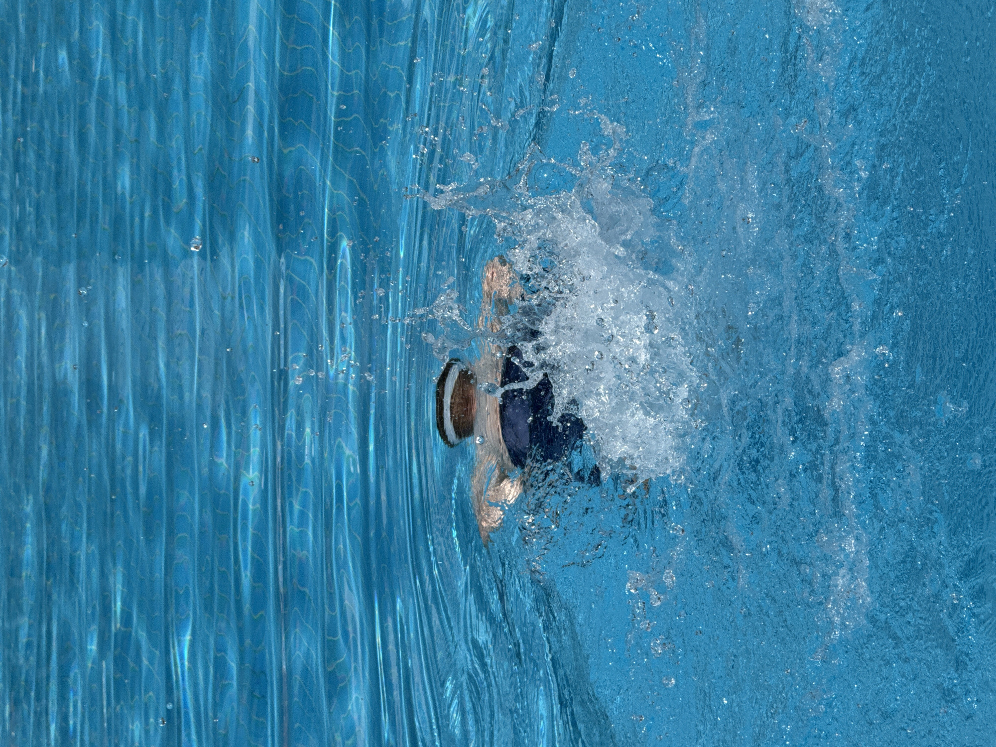 A person swims in a bright blue swimming pool, creating a large splash of water. The swimmer is wearing dark blue swim trunks and a swim cap. The water is clear and reflects the light, creating patterns on the surface. The image appears to be taken from behind the swimmer, looking towards the water. The background consists of more rippling blue water.