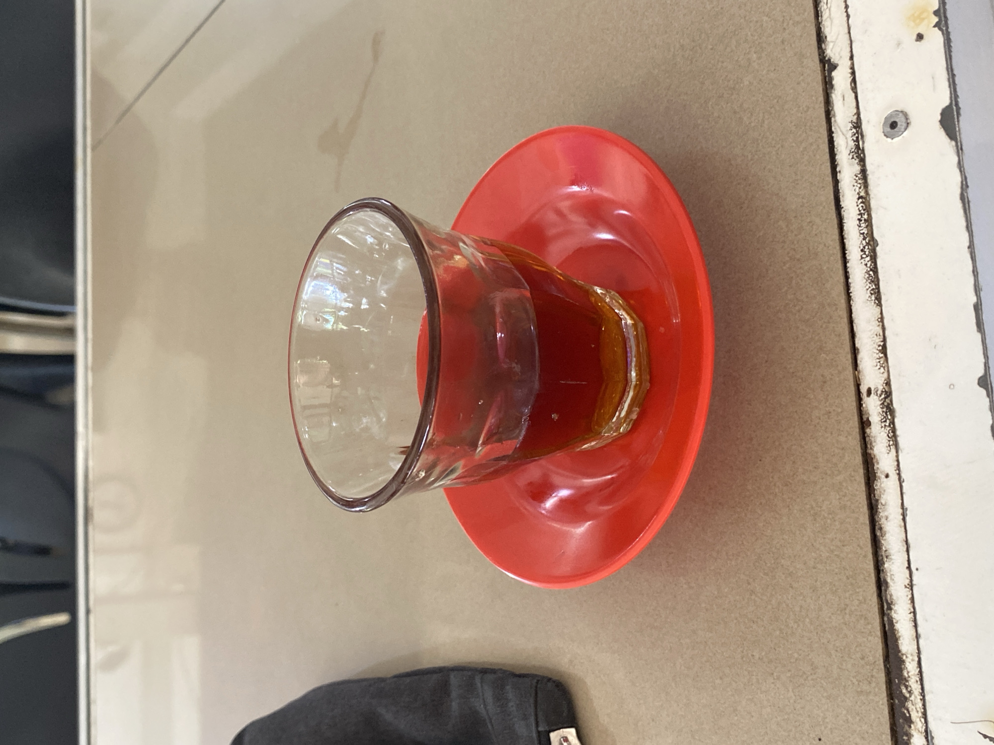 A close-up shot focuses on a clear glass filled with a dark liquid, likely tea or coffee, resting on a vibrant red saucer. The glass has a textured, ribbed design. The scene is set on a light brown, speckled surface that appears to be a table. To the left of the glass, a dark gray cloth item is partially visible, possibly a piece of clothing or a bag. In the background, out of focus, are dark chairs and a white horizontal line suggesting a table edge or a partition. The lighting is bright, creating reflections on the glass and saucer. The setting is consistent with a cafe or restaurant in Lhokseumawe, Indonesia.