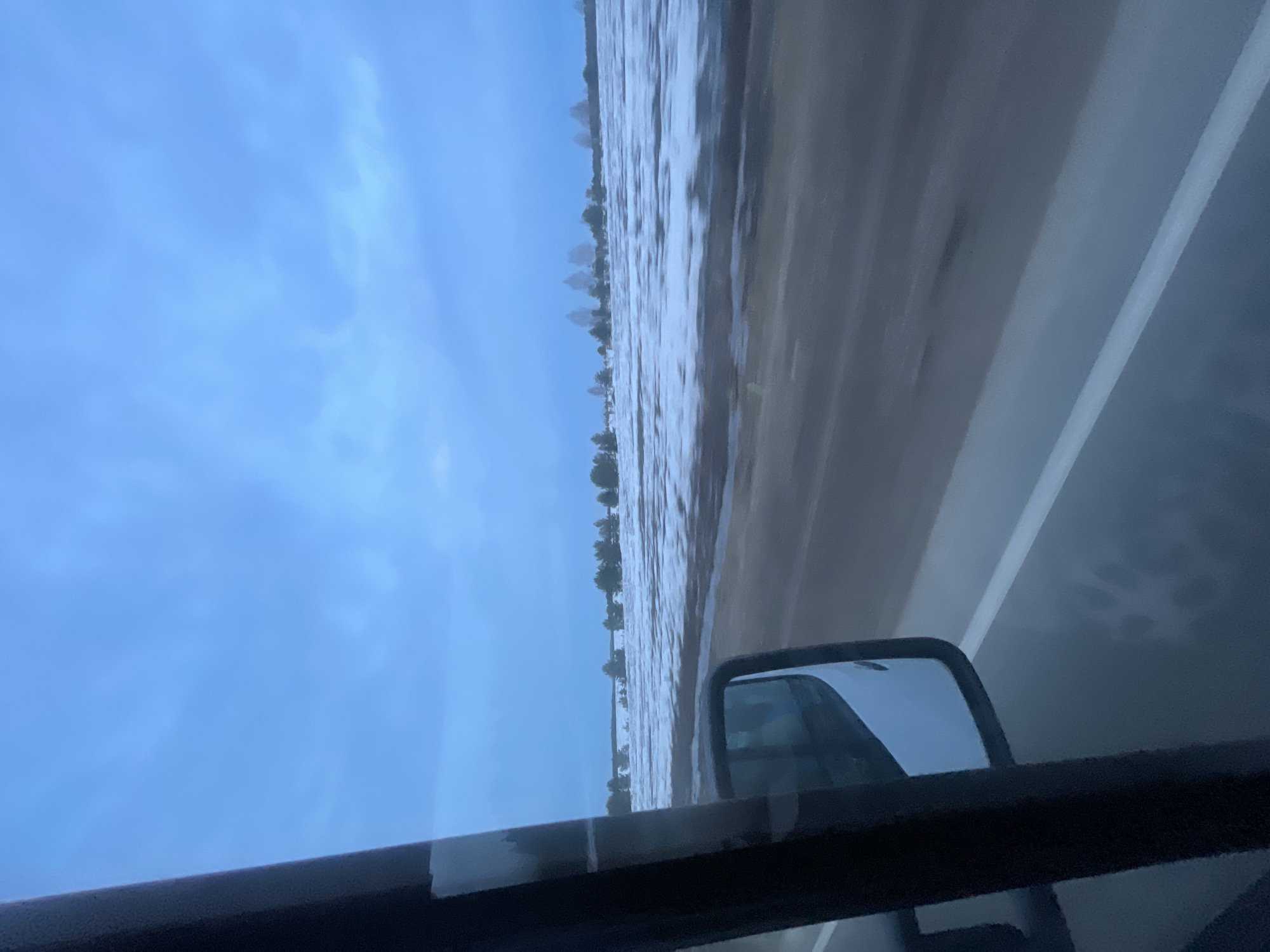 The image, likely taken from inside a vehicle, shows a flooded landscape alongside a road. The road itself is partially submerged, with water reaching up to what appears to be a line of trees or vegetation in the distance. A rearview mirror is visible within the vehicle. The sky is overcast with a blue hue. The reflection visible in the rearview mirror does not provide clear details. The location of the photo is in the city of Povarenki, Russia.
