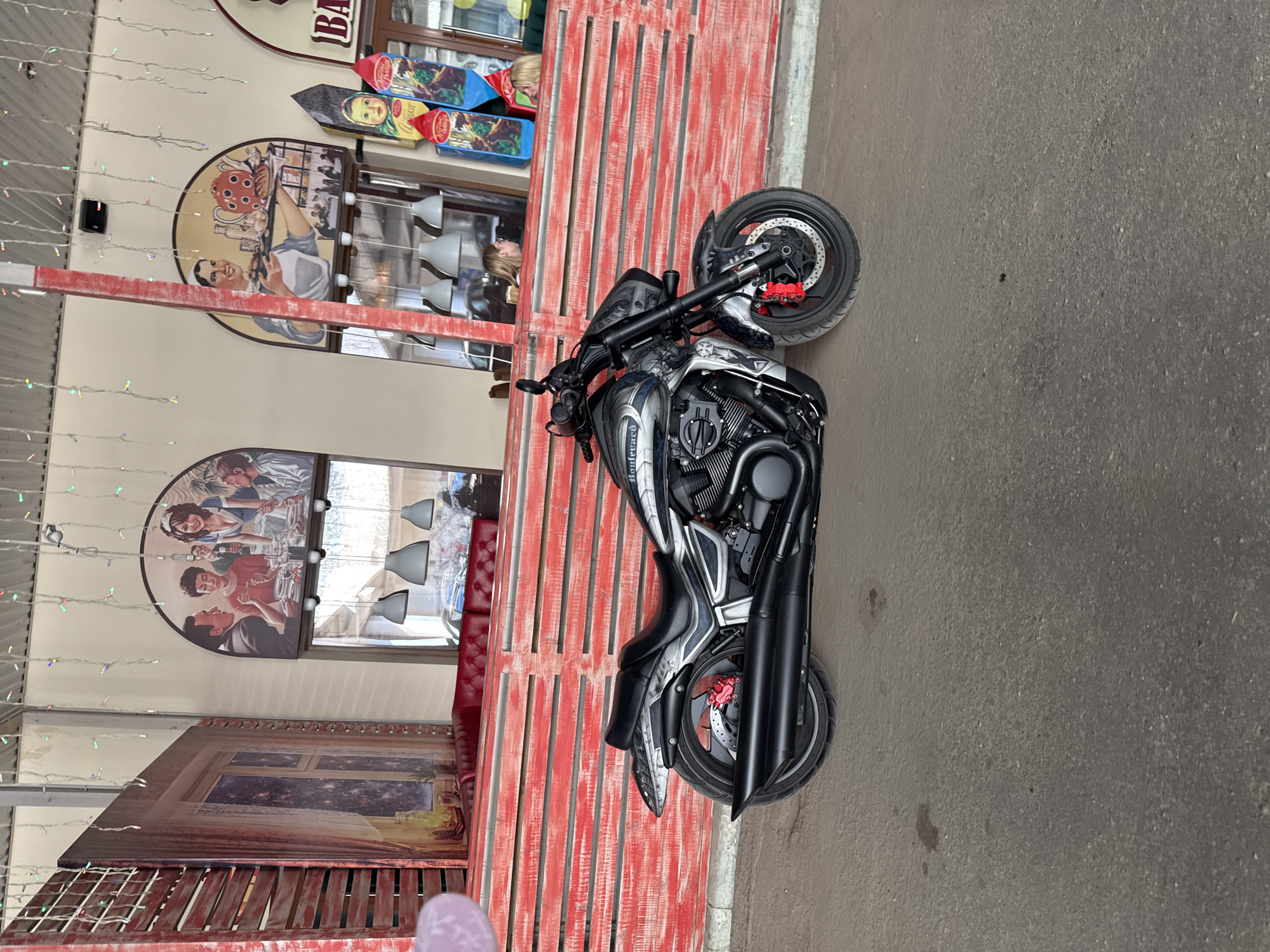 A silver and black Victory motorcycle is parked on a grey asphalt surface, positioned in front of a building with a red wooden facade. The building displays decorative elements, including arched picture frames on the beige wall and string lights hanging from the roof.