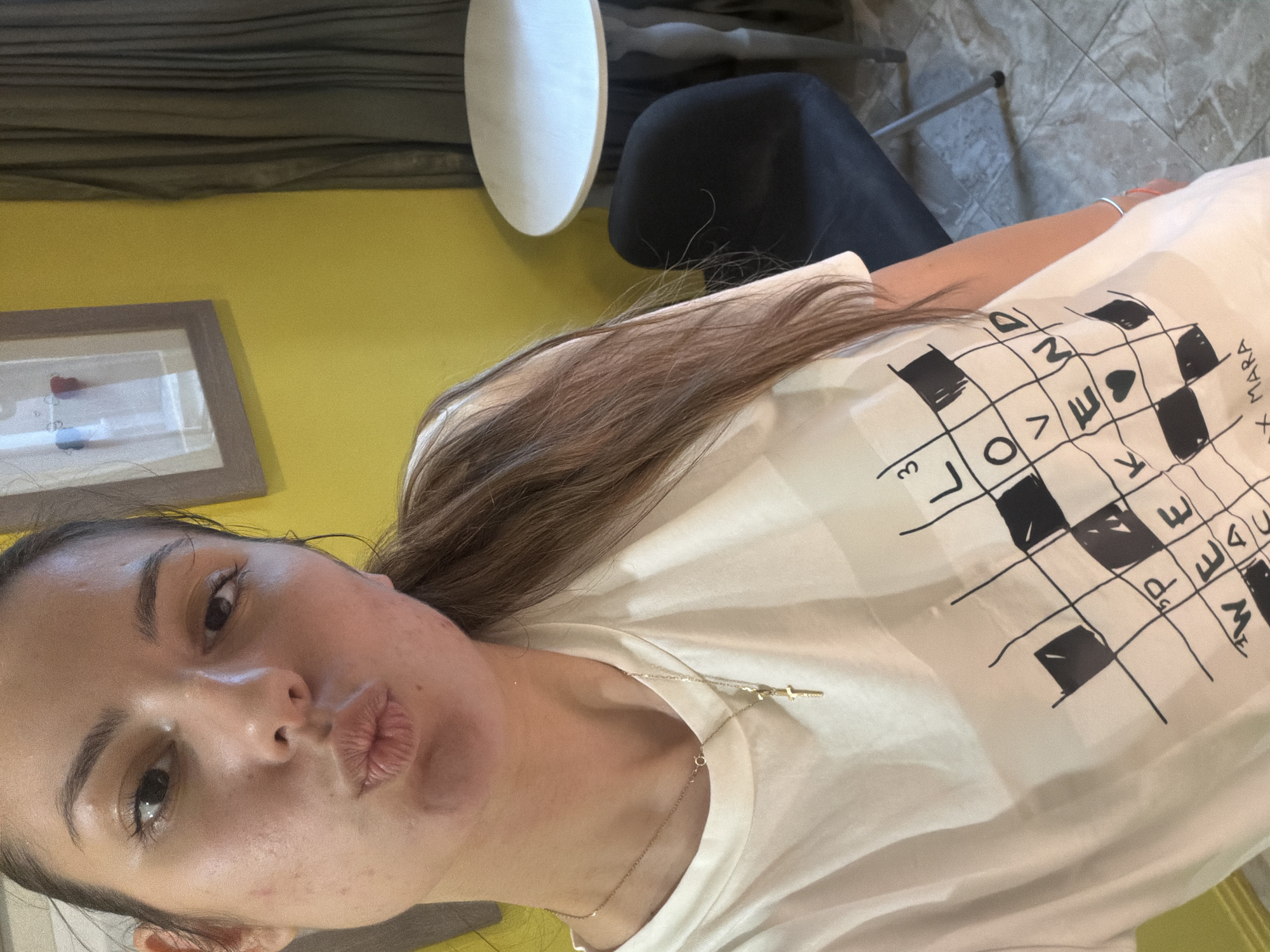 The image displays a selfie of a young woman taken indoors, likely in Burgas, Bulgaria. She is captured with her head tilted, looking directly at the camera with a pouty expression. Her long, dark brown hair cascades over her shoulders. She is wearing a light-colored t-shirt featuring a prominent black grid pattern, resembling a crossword puzzle or a word game board. Several letters are visible within the squares, including "L O V E," "M A R A," and partial letters like "D," "E," "M," and the number "3." A delicate gold chain with a small cross pendant hangs around her neck, and a light blue bracelet adorns her right wrist.
The background shows a vibrant yellow wall. To the left, a framed piece of art or decor, containing small red and blue heart-shaped objects, is partially visible. To the right, a white round table, a black chair, and a tiled floor can be seen, suggesting a casual indoor setting such as a home or cafe. Above the yellow wall, a dark-toned curtain or blind is present. The lighting in the image suggests it was taken during the daytime.