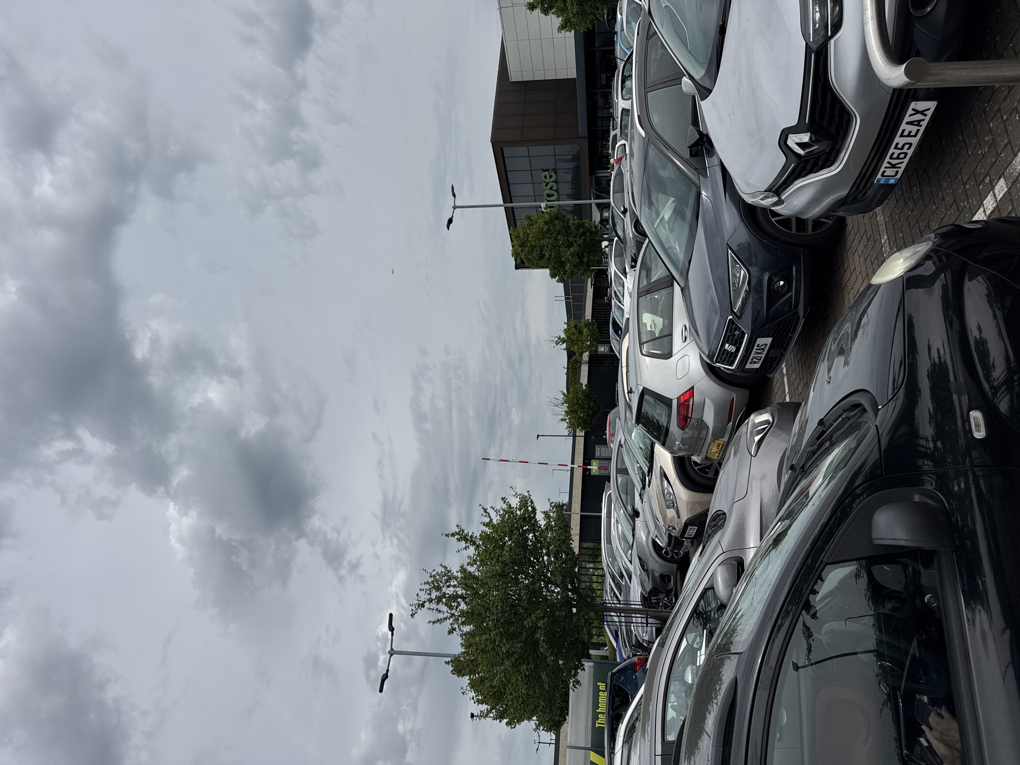 The image shows a car park adjacent to a large building, likely a retail store, in High Wycombe, United Kingdom. Several cars are parked closely together. The sky is overcast with grey clouds. In the background, a tree and a barrier are visible. The building is multi-story, with a glass facade bearing the word "Waitrose". Street lamps are visible throughout the car park.