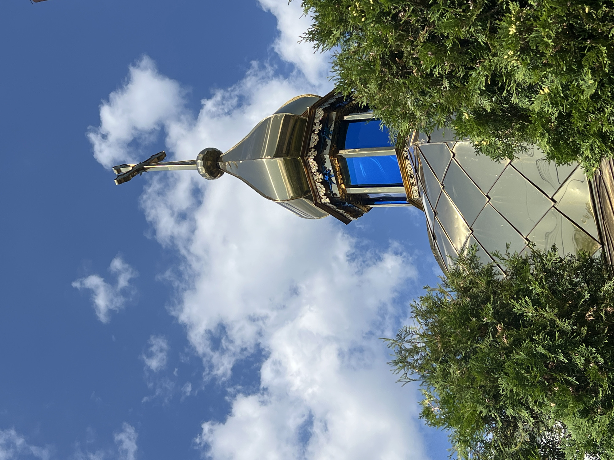 This is a low-angle, outdoor, close-up shot of the golden dome and cross of a church, partially obscured by green evergreen trees. The dome is made of reflective, diamond-shaped panels, and the cross is positioned at the apex of the dome. The sky is bright blue with scattered white clouds. The church appears to be located in Yaremche, Ukraine, a city known for its mountainous scenery and religious sites. There are no people visible in the image.