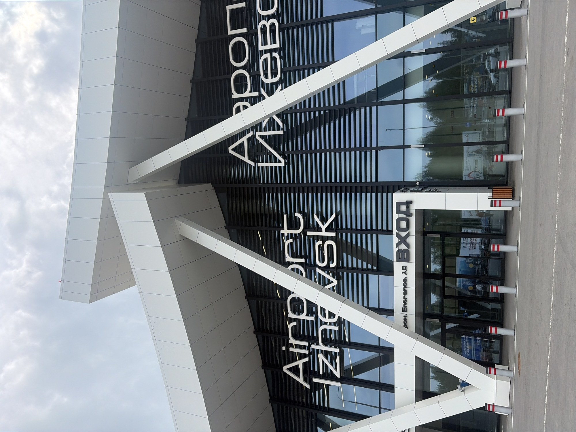 A modern airport terminal building is captured from an exterior perspective, facing its main glass facade. The building features a striking contemporary design with large expanses of reflective dark glass and angular white structural elements made of light-colored panels. These white elements create prominent diagonal lines across the glass front.
Large white lettering is affixed to the glass facade, clearly identifying the building as "Airport Izhevsk" in English and "Аэропорт Ижевск" in Russian. Below these primary signs, smaller white text reads "ВХОД" (Russian for "Entrance") and "Entrance."
The sky above is cloudy, suggesting an overcast or partly cloudy daytime setting. The glass facade reflects the surroundings, including portions of the sky, distant trees, and what appear to be reflections of cars or vehicles on the tarmac.
In the foreground, a paved ground area, likely asphalt, stretches out from the building. Several tall, cylindrical bollards with distinctive red and white reflective stripes are evenly spaced along the edge of this paved area, presumably to guide traffic or pedestrians. The overall scene depicts a clean and well-maintained entrance to a transportation hub, located in Новое Мартьяново, Russia.