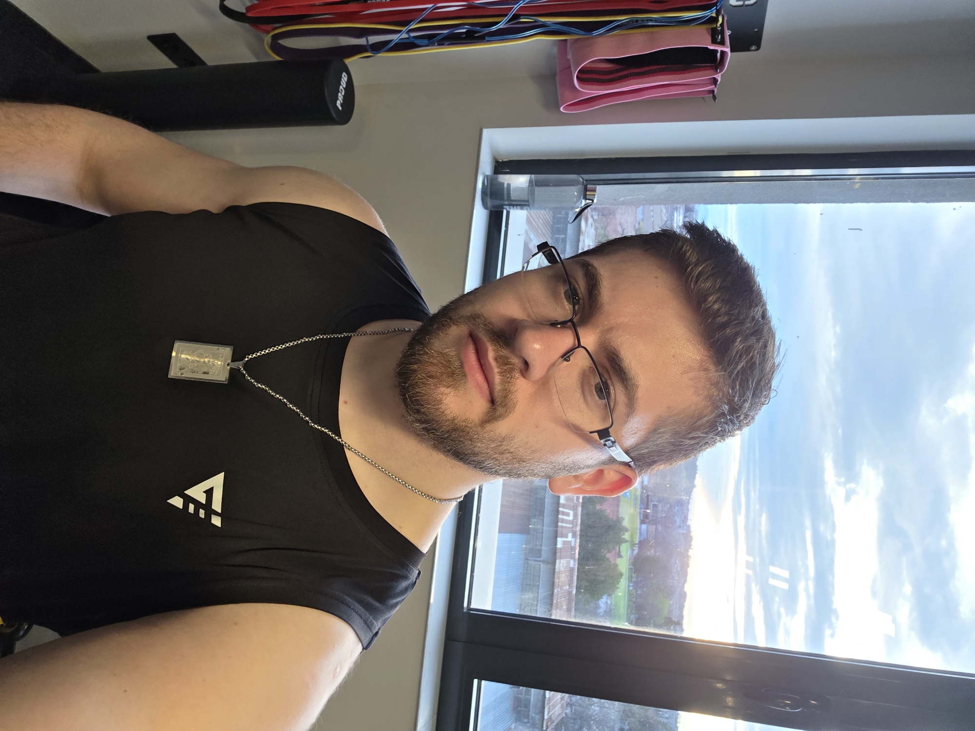 A medium shot depicts a man with a beard wearing glasses and a black athletic shirt with a white logo. He also has a silver chain necklace and dog tags. He is positioned indoors next to a window overlooking an urban landscape. Buildings and greenery are visible through the window, suggesting a city setting. Gym equipment such as exercise bands are mounted on a wall in the background. The exact location within Plovdiv, Bulgaria, cannot be determined from the information in the photo.