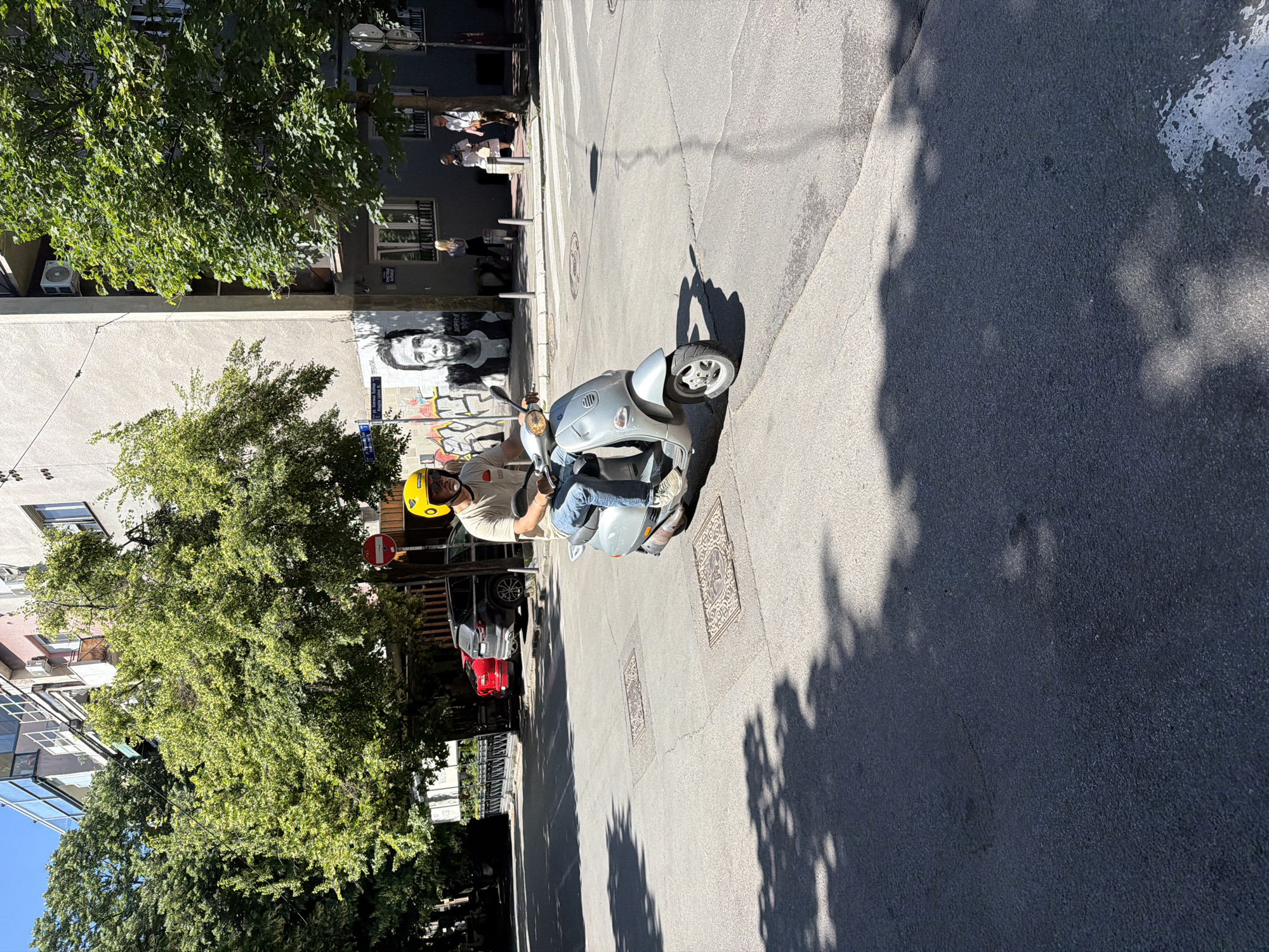 The image depicts a street scene in Belgrade, Serbia, featuring a man on a silver scooter. The man is wearing a yellow helmet, light-colored shirt, and blue jeans. He appears to be stopped on a crosswalk, possibly waiting to cross the street. The street is paved and shows signs of wear. There are trees lining the sidewalk, casting shadows onto the road.
In the background, there is a building with graffiti, including a portrait of a man. The streetscape includes other pedestrians and parked cars. The presence of graffiti and the architectural style are typical of urban areas in Belgrade.