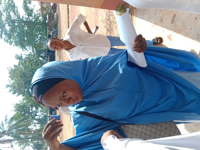 A woman in a blue hijab stands in the foreground, looking down and gesturing with her left hand. She carries a patterned handbag on her shoulder. Behind her, another woman in a white shirt and blue pants stands with a hand raised and a rolled-up paper or document in her other hand. They appear to be outdoors, possibly near a building with trees visible in the background.