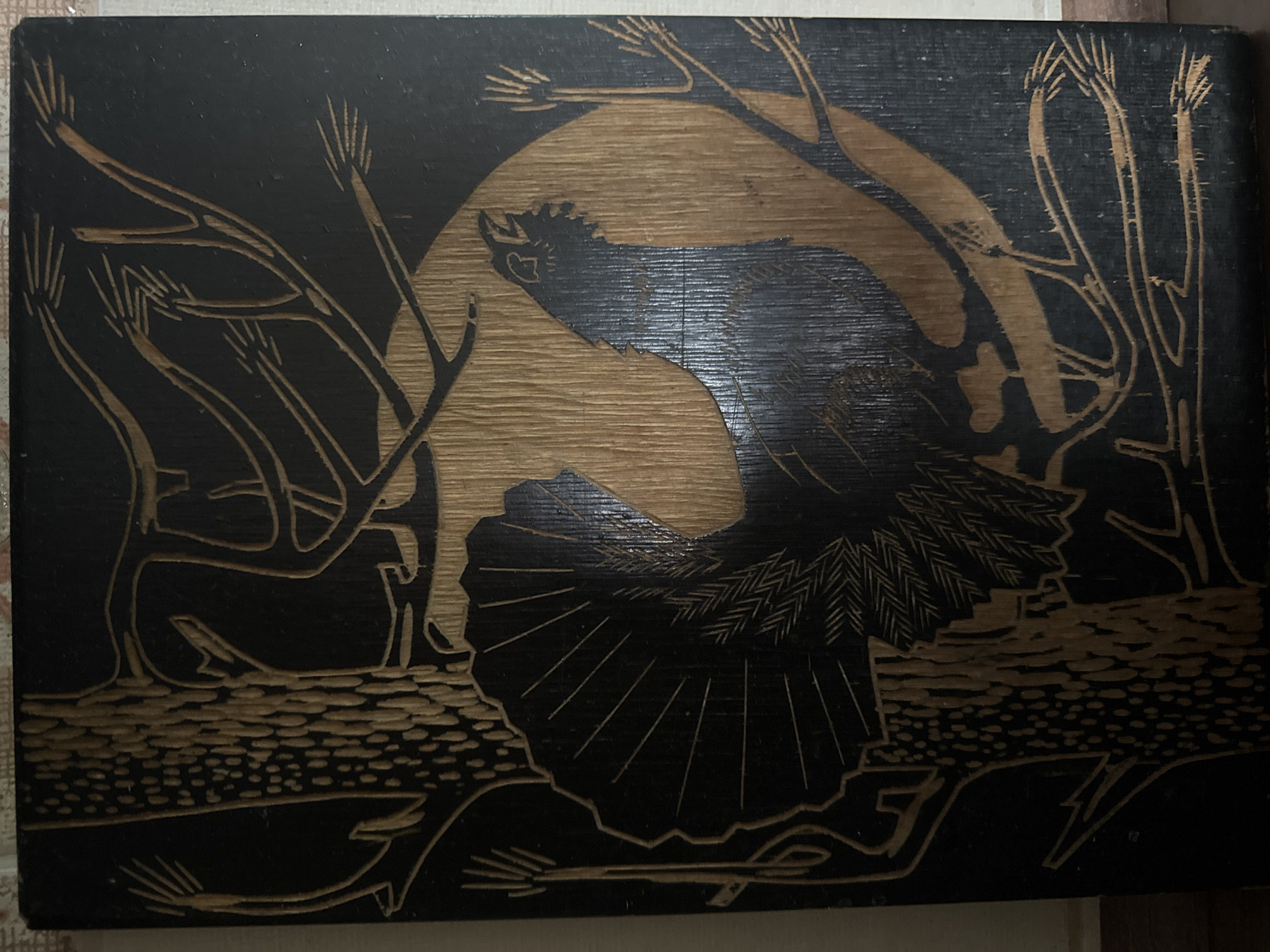 This is a detailed, close-up image of a dark wooden plaque featuring a carved relief of a bird, possibly a capercaillie, with its tail feathers fanned out. The bird is depicted in profile, facing right, with its beak open and head tilted slightly upwards. It is positioned against a large, circular, wood-grain background that represents a moon or sun. Surrounding the bird and moon are stylized branches and pine needles, carved to reveal the lighter wood beneath the dark surface. The tree trunk to the left of the bird is textured with a pattern of carved dots. The carving style is intricate and showcases a contrast between the dark background and the lighter wood of the carved elements. The image does not provide any information about the city of Qonaev, Kazakhstan.