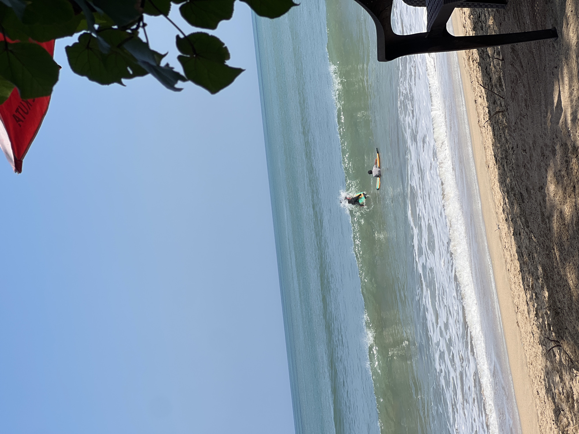 Two surfers are in the ocean off the coast of Kuta, Indonesia. One surfer, wearing a black wetsuit, is actively riding a wave on a colorful surfboard. Another surfer is lying on a yellow surfboard further out in the water. The beach in the foreground is sandy with some debris. A shadow from foliage and a dark chair are visible on the sand. A red umbrella with white lettering, partially obscured by leaves, is at the top of the frame. The sky is clear and blue.