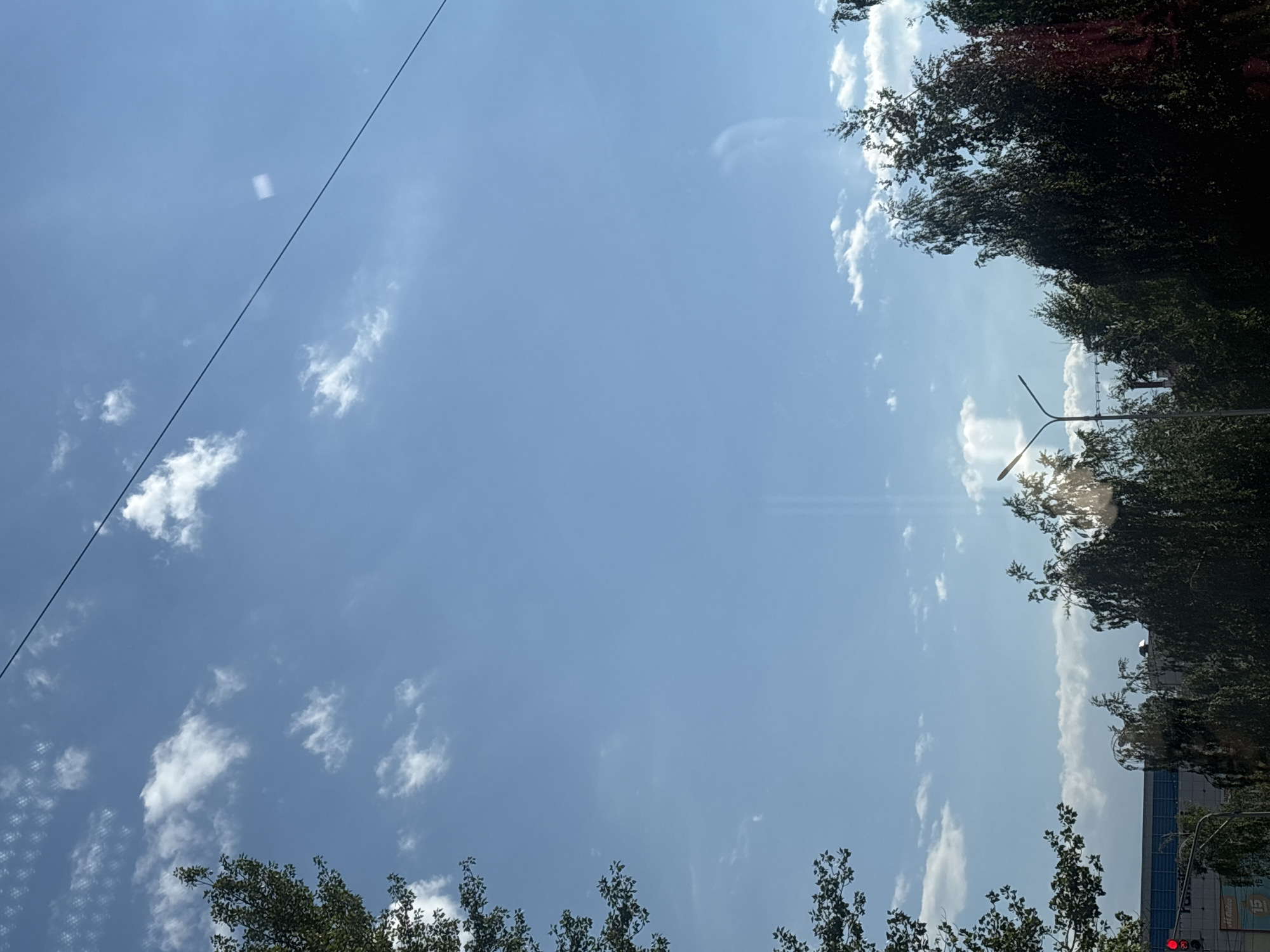 This image depicts a view of the sky and the tops of trees, with hints of urban infrastructure in the lower section, suggesting a location in Ufa, Russia. The sky is a clear blue, populated by scattered white clouds, some of which are wispy and others more defined. A dark line, likely a power line or cable, traverses the upper right portion of the frame. In the lower left quadrant, the branches and leaves of a tree are visible against the sky. The lower portion of the image reveals glimpses of buildings and traffic lights. Specifically, a traffic light with a red illuminated signal is discernible on the far left. Behind the trees, a building with a blue facade and horizontal white lines is partially visible. The overall impression is a bright, daytime scene with a mix of natural elements and urban structures.