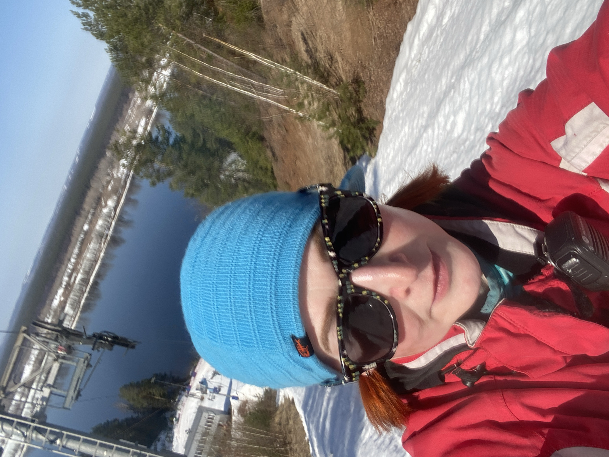 A selfie of a person in a red ski jacket, blue knit hat, and sunglasses, likely taken at a ski resort. The background reveals a snowy slope merging into a landscape with trees and a body of water. A ski lift system is visible in the distance, with structures and cables extending towards the water. Buildings and more trees are also present in the background. The picture appears to have been taken in Прикамский, Russia.
