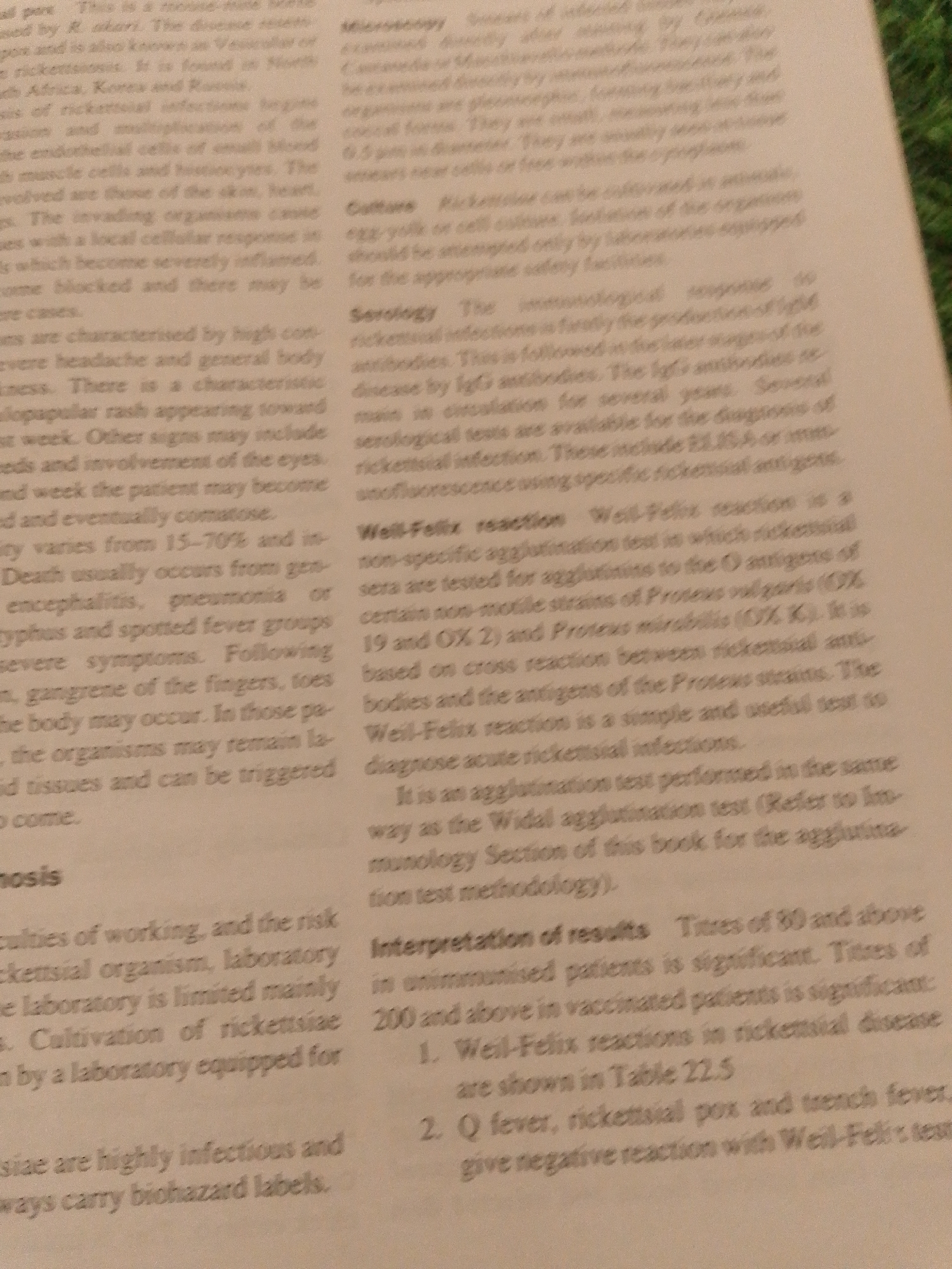 The image is a close-up, slightly angled shot of a page from a medical textbook or document, featuring dense blocks of text. The text discusses rickettsial infections, mentioning microscopy, cultures, serology, and diagnostic tests like the Weil-Felix reaction. It also details symptoms, transmission, and interpretation of results. The lighting is uneven, with a bright light source from the left, creating shadows and making some parts of the text difficult to read. The overall impression is of a scientific or medical reference document. There is no indication of a city, location, or specific setting beyond the printed page itself.