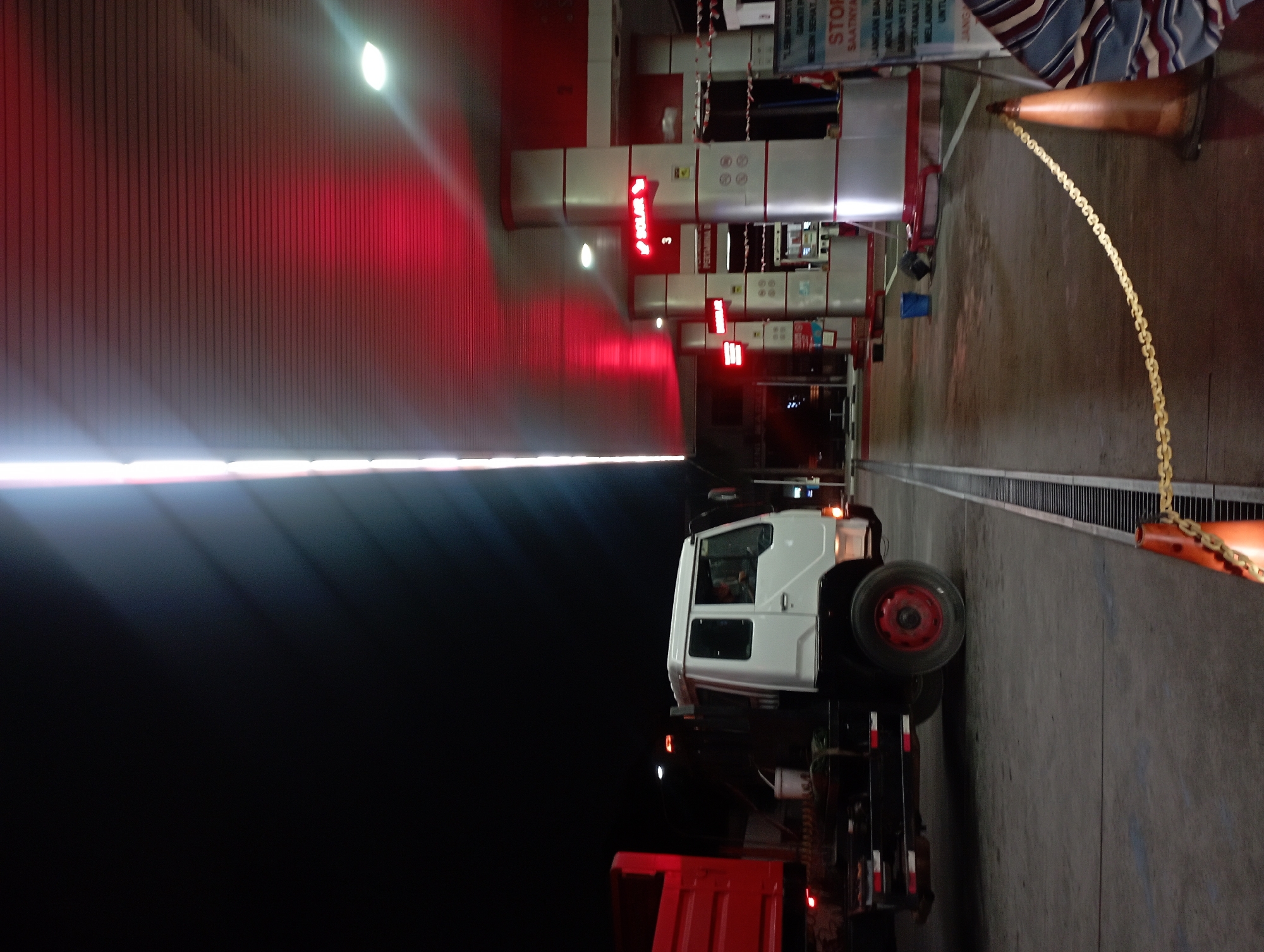 This image captures a night scene at a gas station in Surakarta, Indonesia. A white truck with a red cargo bed is parked on the left side of the frame, its headlights off. The gas station canopy above is illuminated with red lights, casting a vibrant glow. A sign that reads "SOLAR" is visible, indicating the fuel type available. The ground is paved and appears wet in some areas, with a metal grate running along the center of the forecourt. In the background, the gas station's service area is visible, with fuel pumps and a kiosk. A blue bucket and a dark bag are placed on the ground near the pumps. A yellow chain barrier is stretched across the foreground, with an orange traffic cone in the lower right corner. The overall atmosphere is that of a typical, well-lit gas station at night.