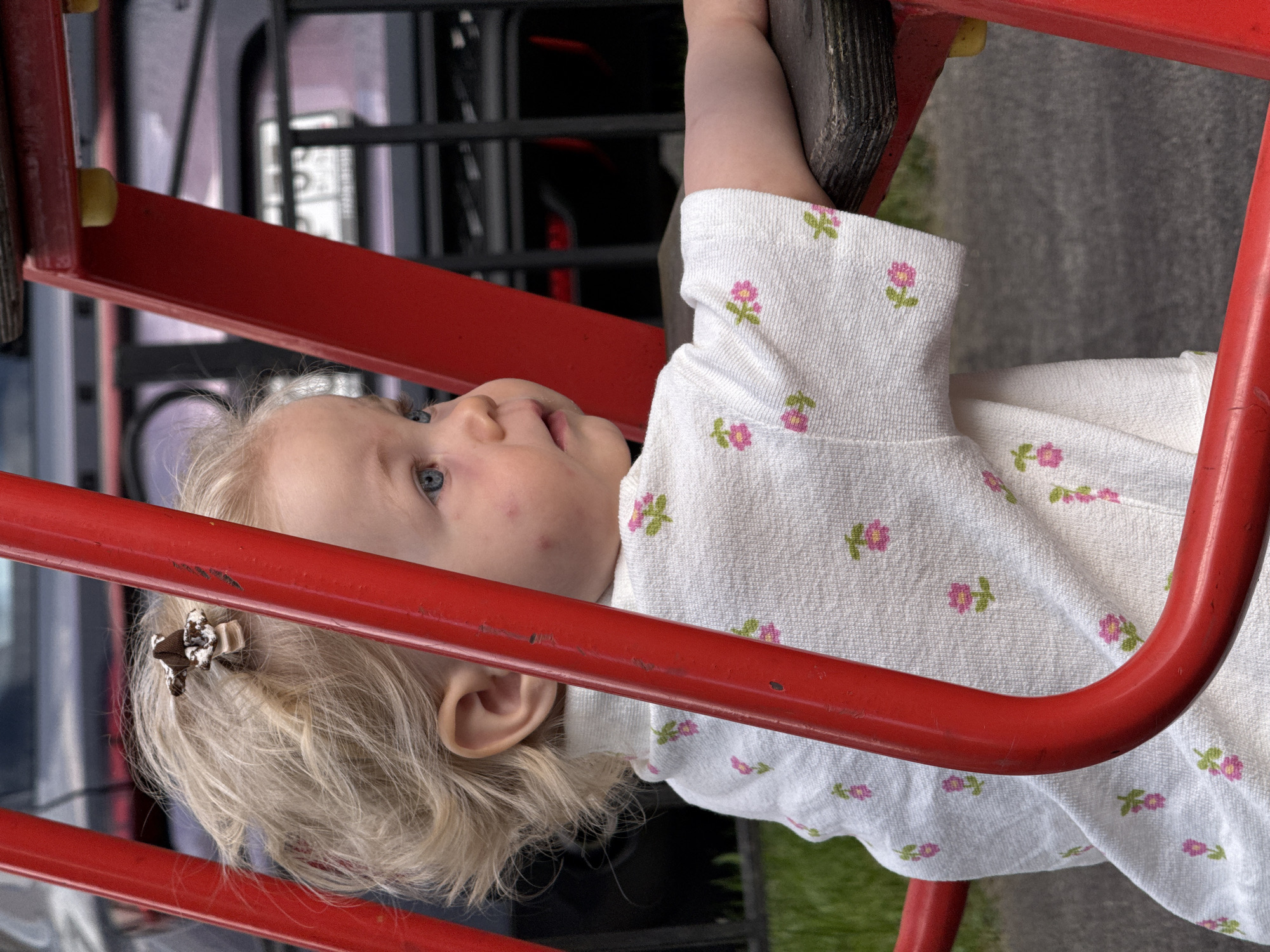 An outdoor, eye-level, medium shot captures a blonde-haired toddler with blue eyes and light skin, wearing a white shirt with a floral pattern of pink flowers and green leaves. The child is positioned in profile, looking to the right, with a small brown clip in their hair. The toddler is leaning on a wooden platform, which is part of a red metal structure, possibly a playground. The background is blurred but suggests an urban environment with a dark vehicle and some greenery. The lighting is natural and appears to be overcast. There are no visible landmarks to pinpoint the location in the city of Vladimir, Russia.