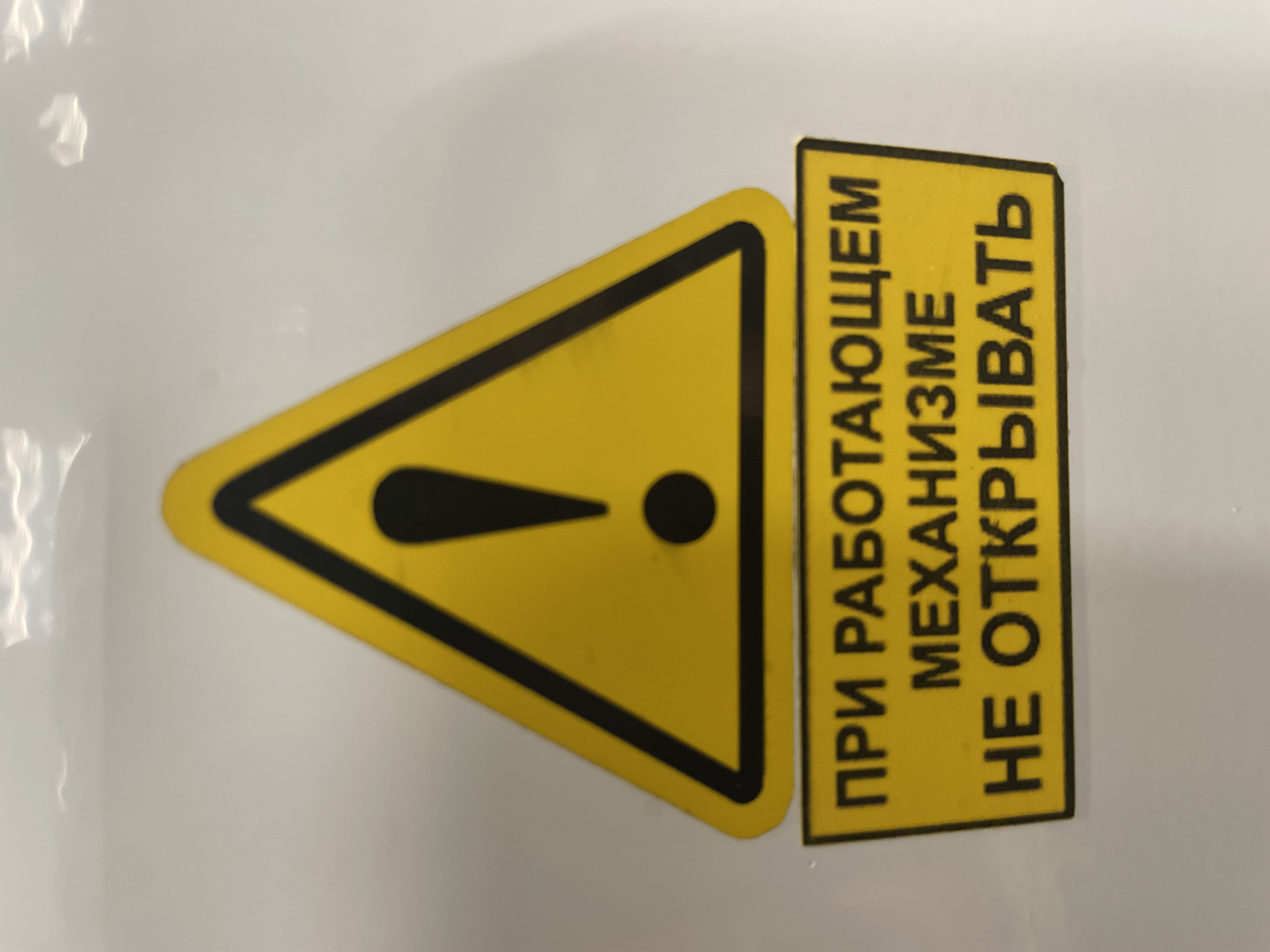 The image shows a yellow triangular warning sign with a black border and a black exclamation mark in the center. Below the sign, there is a yellow rectangular label with black text in Russian. The text reads: "ПРИ РАБОТАЮЩЕМ МЕХАНИЗМЕ НЕ ОТКРЫВАТЬ", which translates to "DO NOT OPEN WHILE THE MECHANISM IS RUNNING". The sign is affixed to a white surface, which appears to be a wall or a piece of equipment. The background is mostly white with some subtle shadows and reflections. There are no people or animals visible in the image. The image appears to be a close-up shot of the warning sign and label. There is no information in the image that can be used to determine its location in the city of Терволово, Russia.