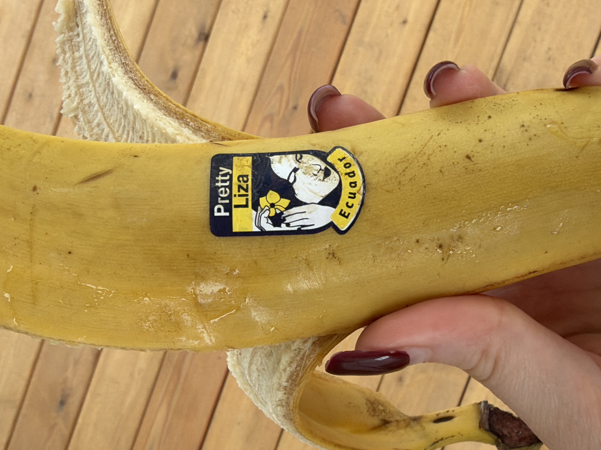 A partially peeled banana is held in a person's hand. The banana has a "Pretty Liza Ecuador" sticker. The background is a light-colored wooden surface.