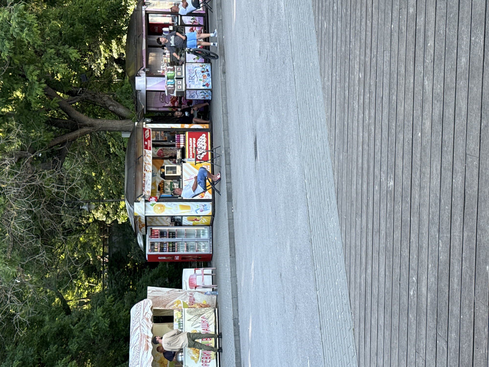 The image depicts a street scene in Varna, Bulgaria, likely a promenade or boardwalk area, with a row of food kiosks set up along a paved walkway. In the foreground, a wooden deck extends across the bottom of the frame. Behind the deck is a grey paved pathway, and then the row of kiosks.
On the left, a kiosk displays signs with Cyrillic text and graphics of food items, including corn. Two men are visible at this kiosk; one is standing and appears to be working, while another is partially visible behind him. A red plastic chair sits next to the kiosk.
To the right of the first kiosk, a red refrigerator unit is stocked with beverages. Adjacent to this is another kiosk with colorful graphics and offerings, including ice cream. A man is seated at a small table in front of this kiosk.
Further to the right, another kiosk features more beverage displays and what appears to be ice cream or gelato. A young man with a bicycle stands in front of this kiosk, dressed in a t-shirt and shorts. A second man is seated at a table to the right of this kiosk.
In the background, lush green trees form a dense canopy, suggesting a park-like setting or the edge of a green space. Sunlight filters through the leaves, creating dappled light. A lamppost is visible among the trees. The overall atmosphere is casual and suggests a warm, possibly summer, day.