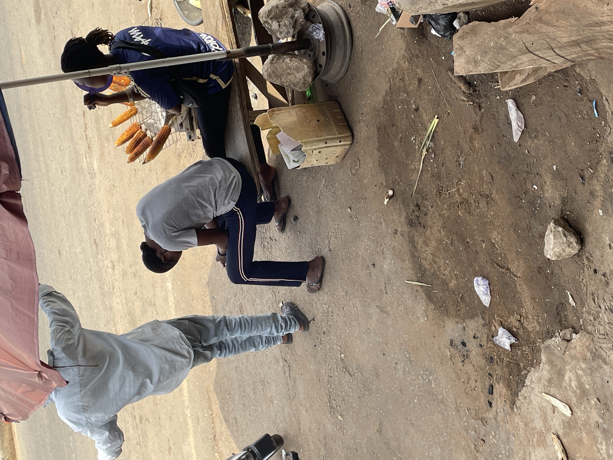 This image depicts a street scene in Akwanga, Nigeria. In the foreground, a person is inspecting roasted corn cobs arranged on a grill. Nearby, another person leans down, possibly examining something on the ground. A third person stands close by, partially obscured by an awning. The background shows a dirt surface, littered with stones and debris. A makeshift structure supports the grill, utilizing found objects such as rocks and a wheel rim.