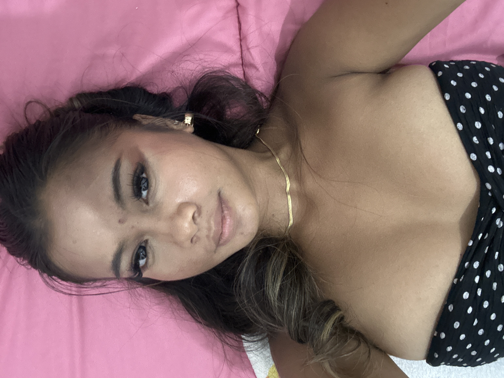 A person is lying on a pink surface. They have dark hair with lighter strands and are wearing a black and white polka dot strapless top. They are also wearing a gold chain necklace and earrings. The location is unknown, and there is no evidence to suggest the city of Celuk, Indonesia.