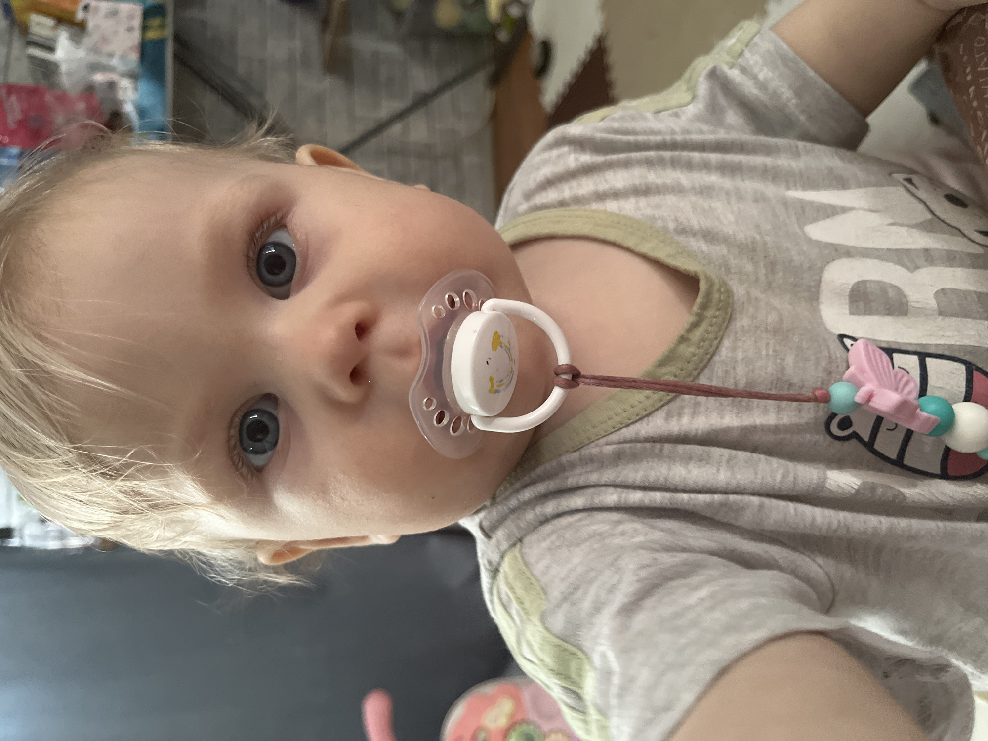 The image presents a close-up, sideways-oriented view of a baby or toddler with fair skin, light blonde hair, and prominent blue eyes, looking directly at the camera. The child has a white pacifier in their mouth, which features a subtle yellow animal graphic, possibly a chick or duck. A pacifier clip, consisting of a brown string, light blue, white, and pink beads, and a pink butterfly-shaped charm, is attached to the pacifier and secured to the baby's t-shirt.
The baby is wearing a light grey t-shirt with light olive-green trim around the collar and sleeves. Visible text on the shirt includes large white letters spelling "BRO", accompanied by a graphic of a cartoon character's face with a wide mouth. The background is blurred, suggesting an indoor setting, possibly a home, with various indistinct objects. The baby's expression appears neutral and curious. No specific time of day, weather, or discernible location cues within the image itself are apparent, though the provided context indicates the location is Vesyoloye, Russia.