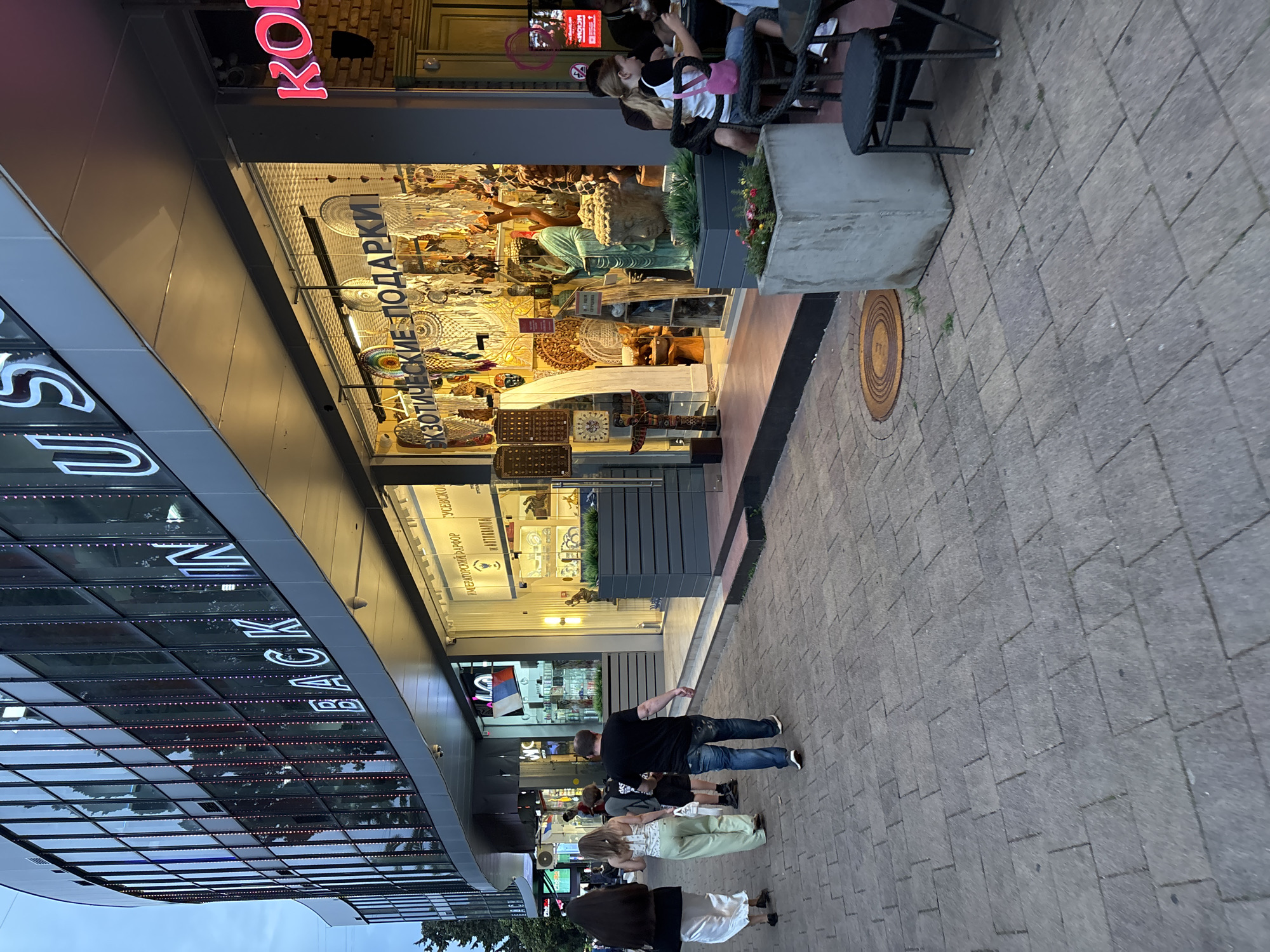 The image depicts a street scene in Sochi, Russia, specifically along a commercial strip. A modern building with large glass windows curves along the street, displaying signs with white illuminated text that reads "BACK IN US". Adjacent to this building is a shop with a prominent window display showcasing a variety of exotic gifts, including dreamcatchers, statuettes, and decorative items. In front of the shop, several people are walking on a paved sidewalk. One person, a man in a black t-shirt and blue jeans, is walking away from the camera. To his left, two women are walking in the same direction, one in a white top and light green pants, the other in a black top and white skirt. Further down the sidewalk, a few individuals are seated at outdoor tables, suggesting a cafe or restaurant area. The overall environment suggests a moderately busy urban setting with commercial establishments and pedestrian activity.