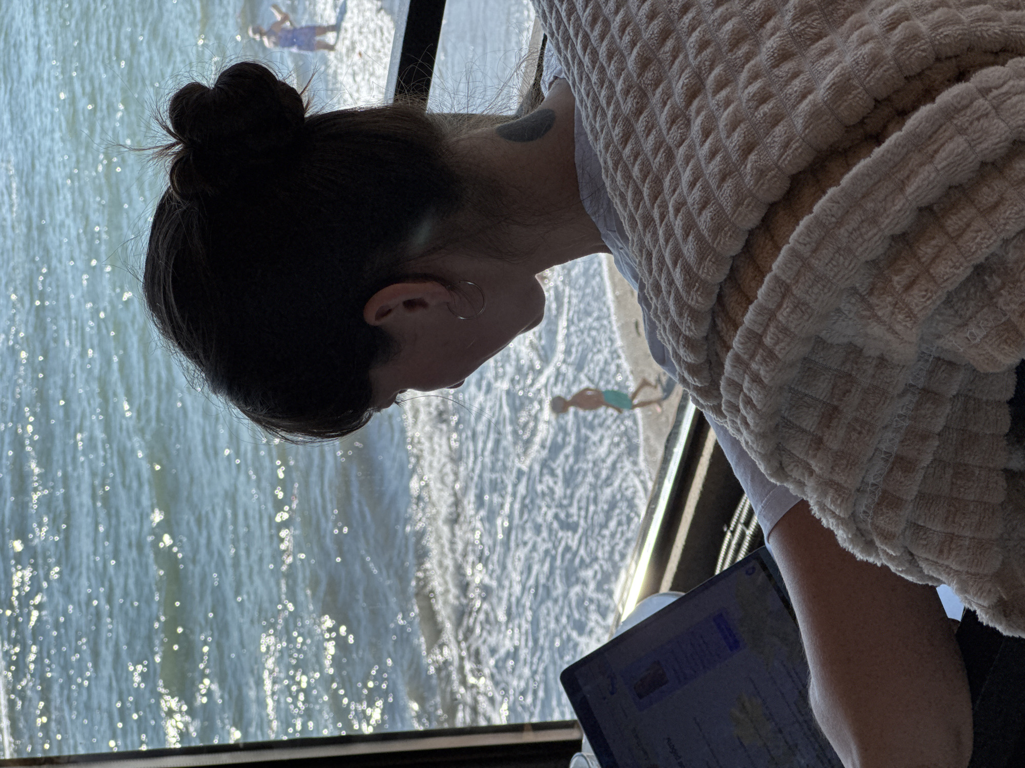 A woman with dark hair styled in a bun, wearing a light-colored t-shirt and a tan, textured blanket draped over her shoulders, sits in front of a laptop. She is positioned with her back to the camera, facing a window that overlooks a beach and the sea. The sea is bright with sun glare, and gentle waves are breaking on the shore. A small child in swim trunks walks along the waterline in the distance. The scene appears to be an indoor cafe or workspace with a view of the Black Sea coast in Sochi, Russia.