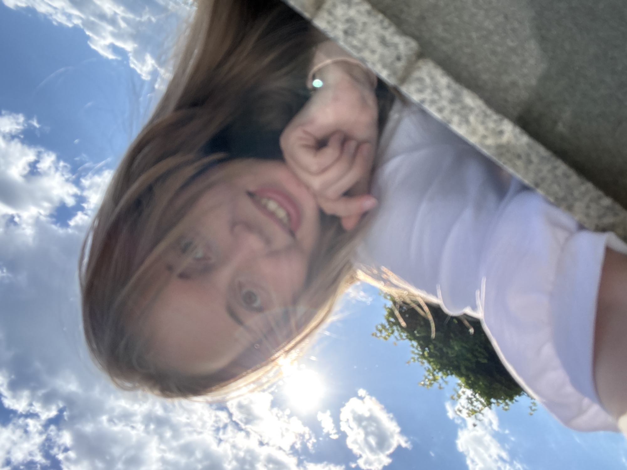 A young, light-skinned woman is depicted outdoors on a sunny day in an unidentified location (Unknown, Unknown). The image is captured from a low-angle perspective, looking upwards towards the sky. The woman, positioned in the center-right of the frame, smiles directly at the camera with her teeth visible. She has long, light brown hair, which is backlit by the sun, creating a prominent lens flare originating from the upper-left quadrant. She is wearing a white long-sleeved top. Her right arm is bent, and her right hand is brought up towards her chin, with fingers slightly curled. A thin, light-colored bracelet with a small, luminous circular bead is visible on her right wrist. The background consists of a bright blue sky filled with numerous white cumulus clouds. A patch of green foliage is visible in the lower-left background. A textured, granular grey surface, resembling a concrete or stone ledge, occupies the bottom-right and right edge of the foreground.