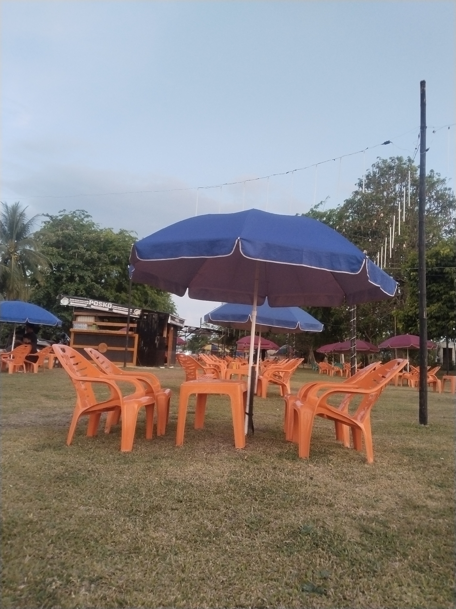 The image shows an outdoor seating area in what appears to be a park or recreational space. Several orange plastic chairs and tables are arranged under blue and maroon umbrellas on a grassy lawn. A small building or kiosk labeled "POSKO" is visible in the background, suggesting a service or information point. Trees surround the area, and a tall pole with hanging decorations stands in the background. The sky is light blue, indicating either early morning or late afternoon light. No people are visible. The location, though indicated to be Glokaron, Indonesia, lacks specific landmarks to confirm.